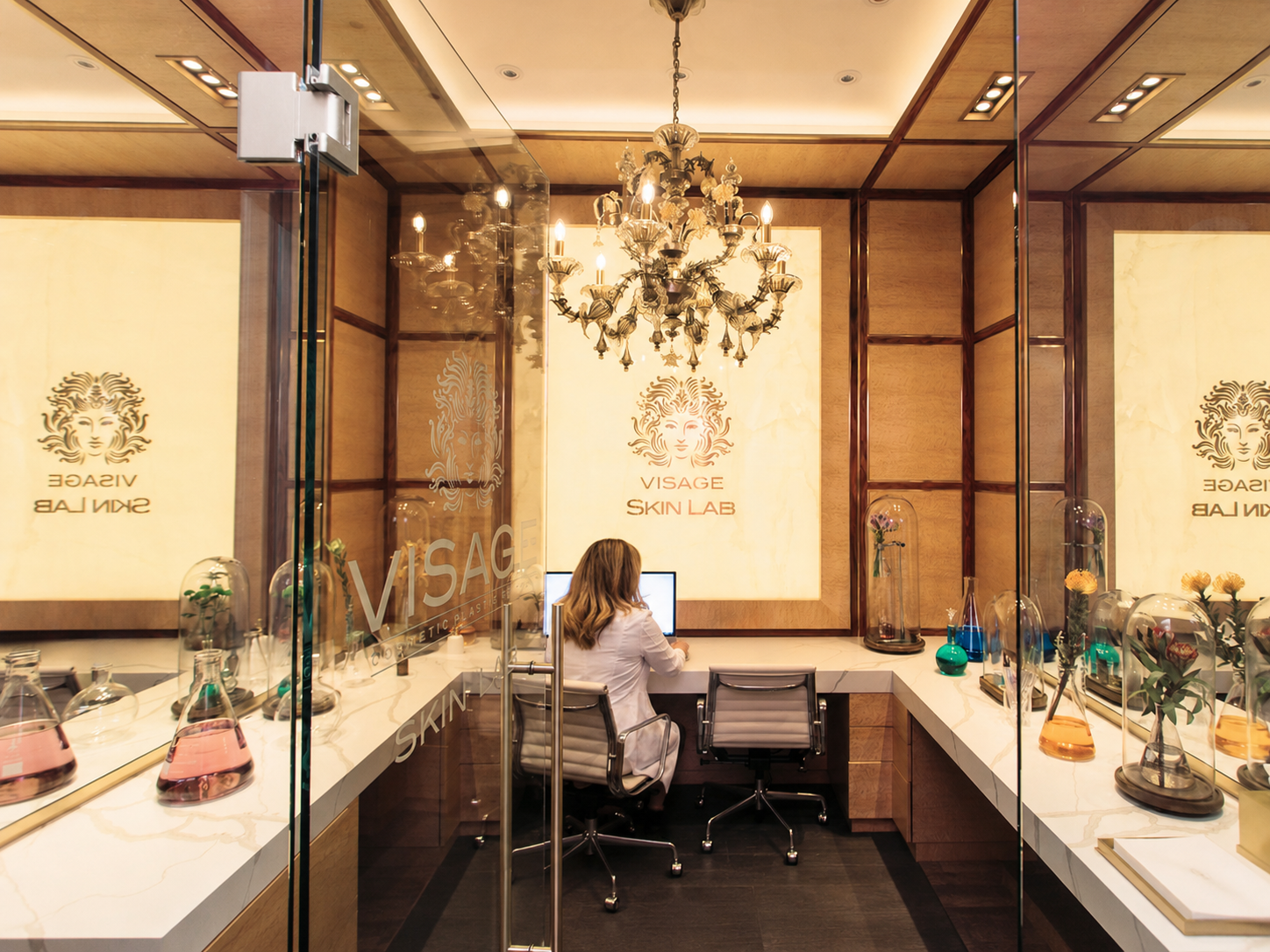 Interior of a skincare clinic with a chandelier and glass display cases filled with bottles and flowers, featuring warm lighting and wood paneling on the walls.
