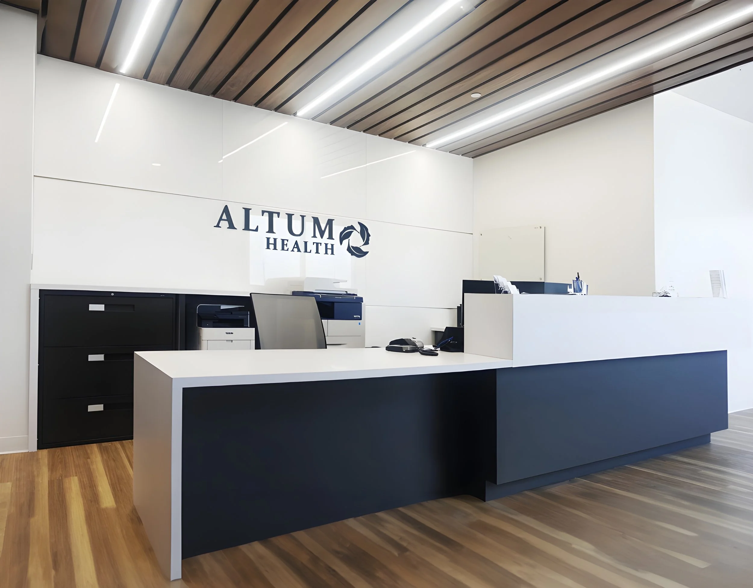 Reception area with white walls, a wooden ceiling with linear lights, and a black and white reception desk with computers and office supplies.