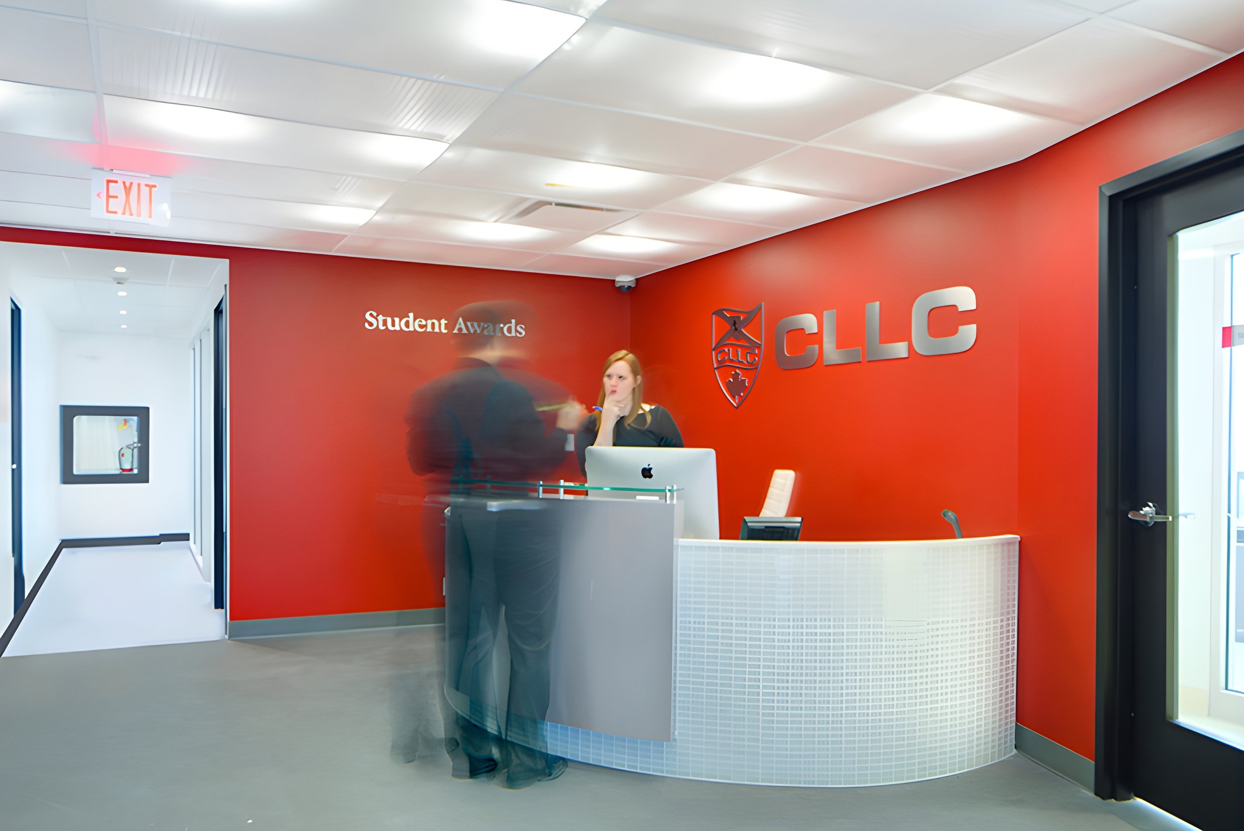 Reception area with a curved white desk, a woman sitting behind it, and a man standing in front of her. The background features a red wall with the logo 'CLLC' and the words 'Student Awards'. A doorway leads to a corridor on the left, and there is an
