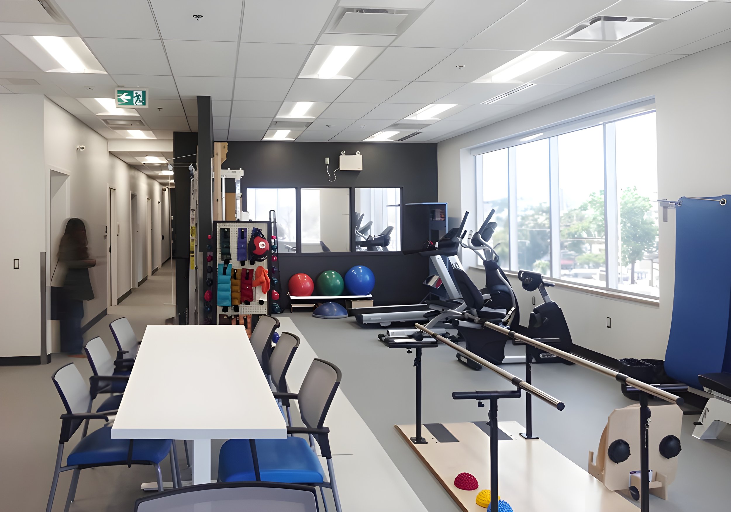 A room with exercise equipment, chairs around a table, and large windows letting in natural light.
