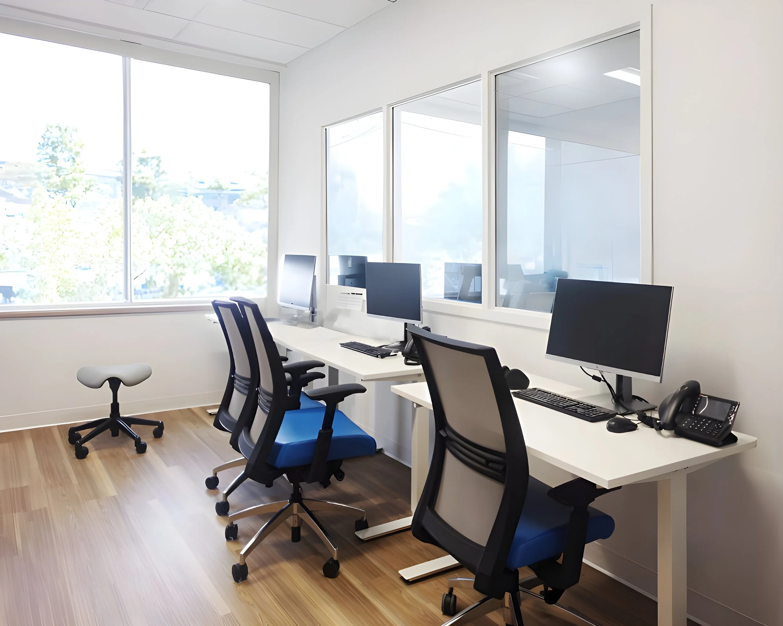 Office room with large window, multiple computer desks, black and blue office chairs, and office equipment including monitors and phones.