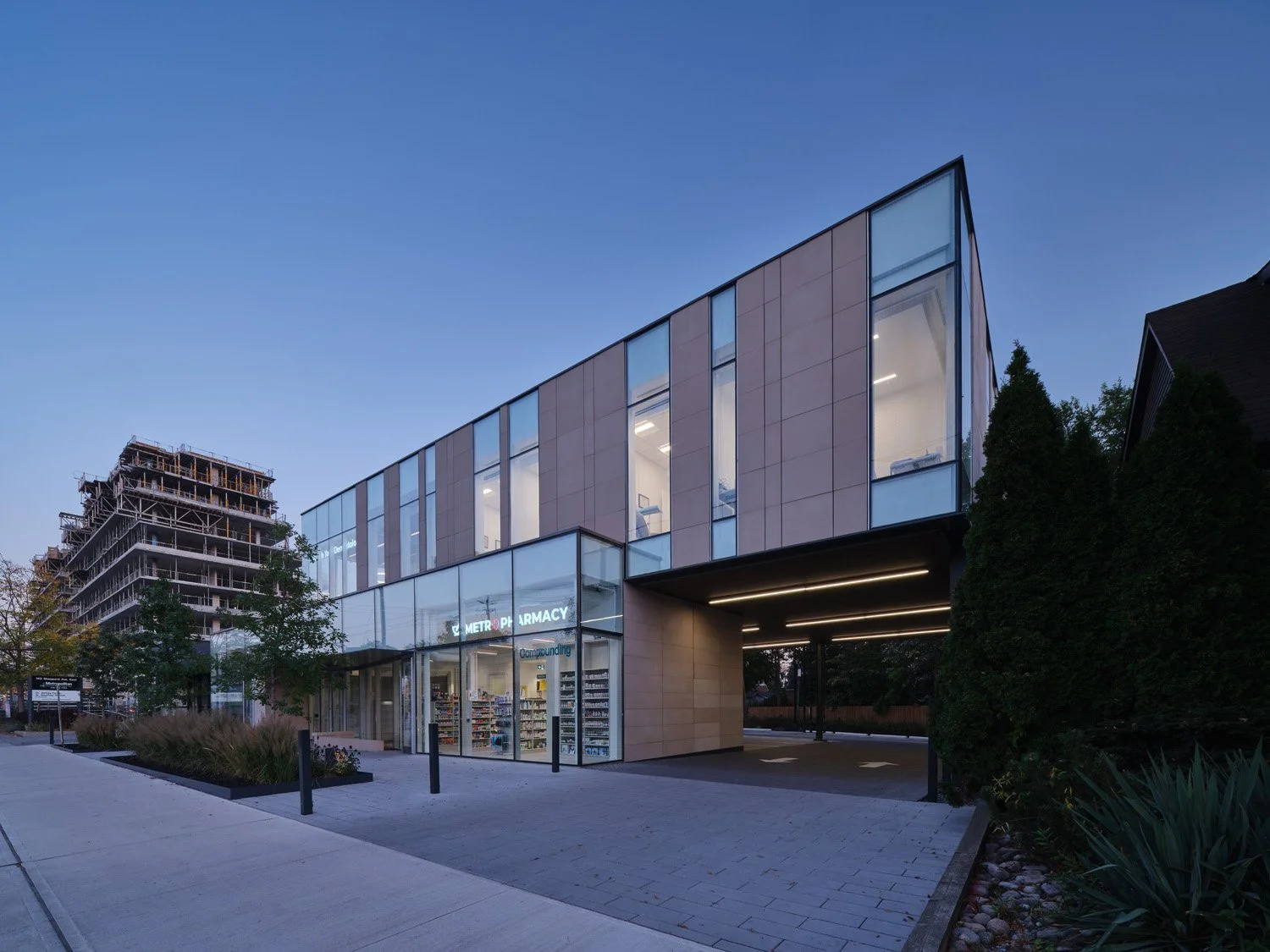 Modern two-story building with glass windows and a pharmacy on the ground floor, surrounded by trees and shrubbery, during dusk. Salutogenic and healthcare design. Good flow and wayfinding.  Good inspiration for Toronto, Dallas and Park City.