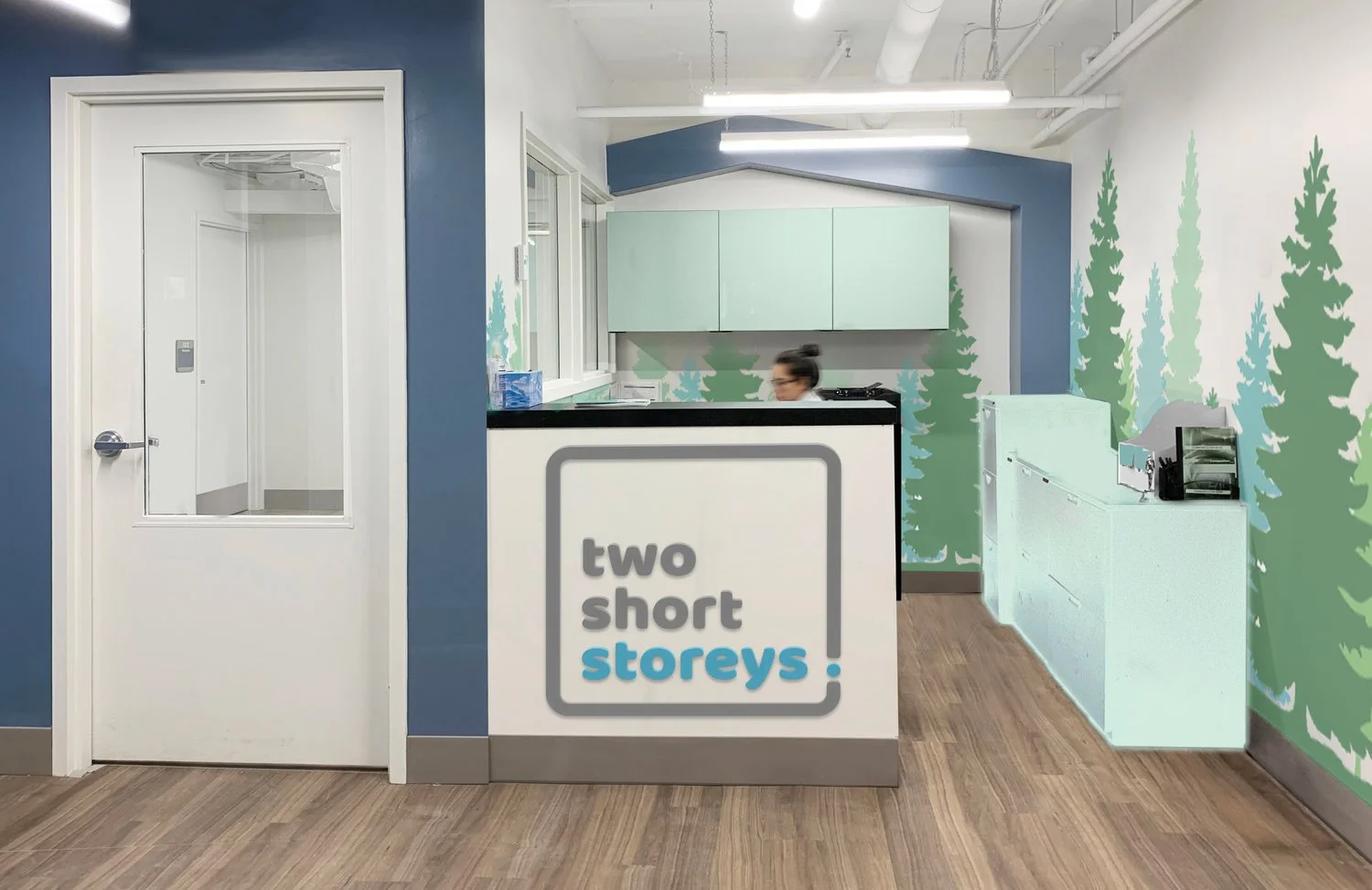 Reception area with a sign that says "two short storeys" in front of a woman standing behind a counter. The background wall has a tree mural with green and blue trees painted on a white wall.