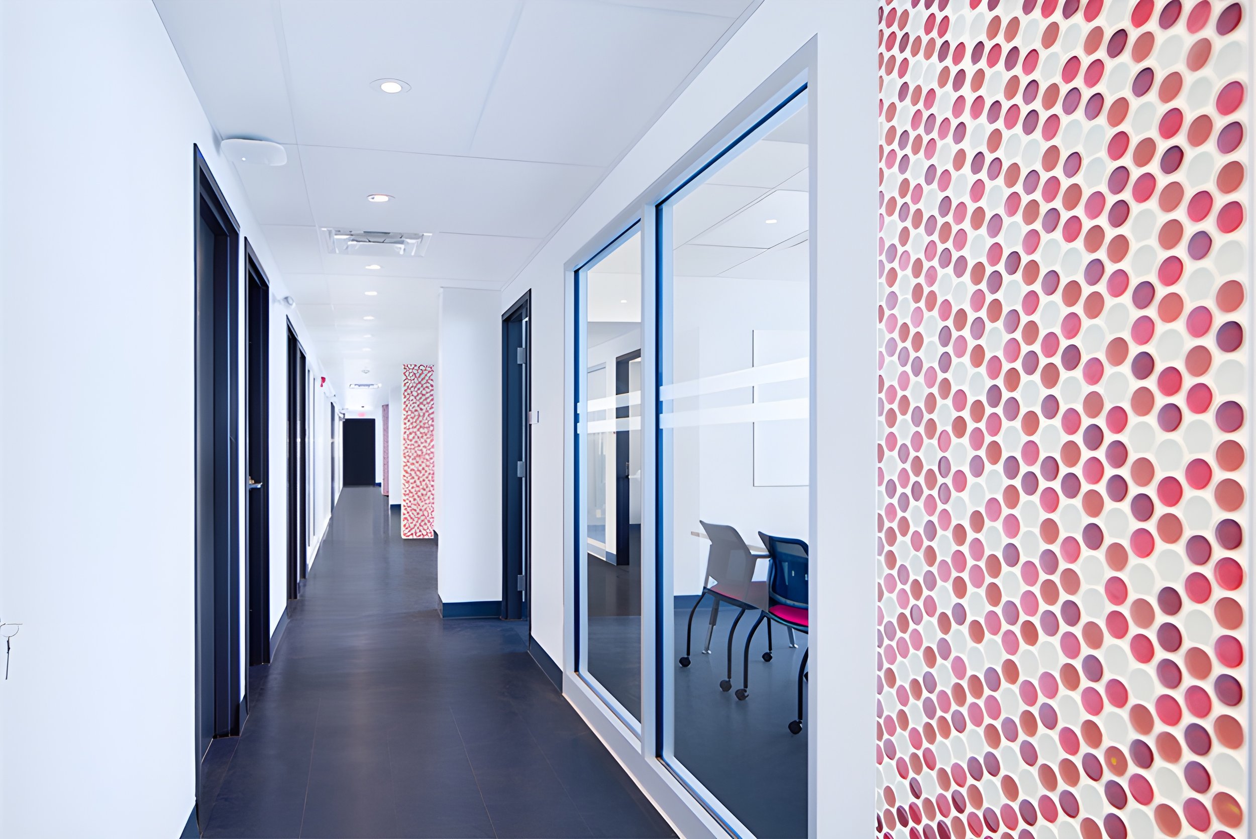 Modern office corridor with white walls, dark flooring, glass-walled conference rooms, and a decorative wall with red and white circular patterns.