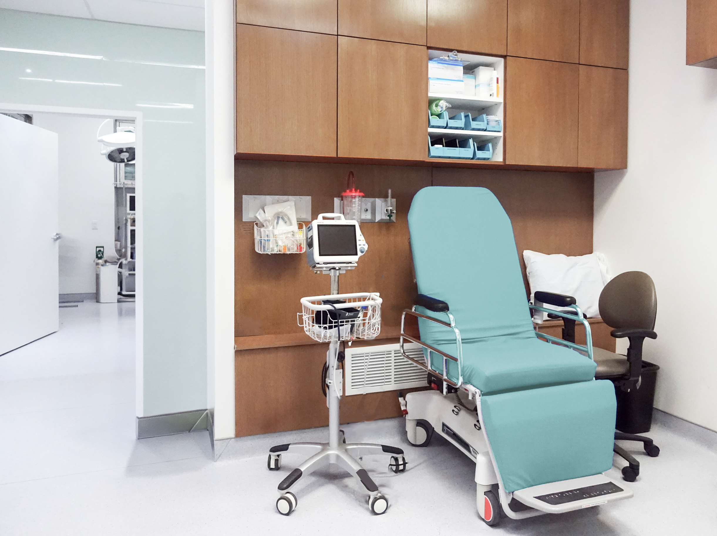 Hospital room with a teal treatment chair, medical equipment, and wall-mounted wooden cabinets.  healthcare, wellbeing. salutogenic design. wayfinding, human. inspiration for Toronto, Dallas, Texas, Miami, and Park City.