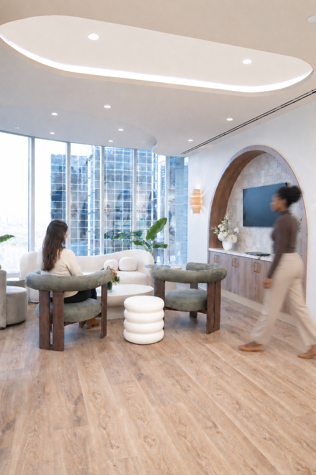 Modern living room with large windows, white sofa, green chairs, wooden floor, and a flat-screen TV mounted on a wall with decorative flowers and lamps.