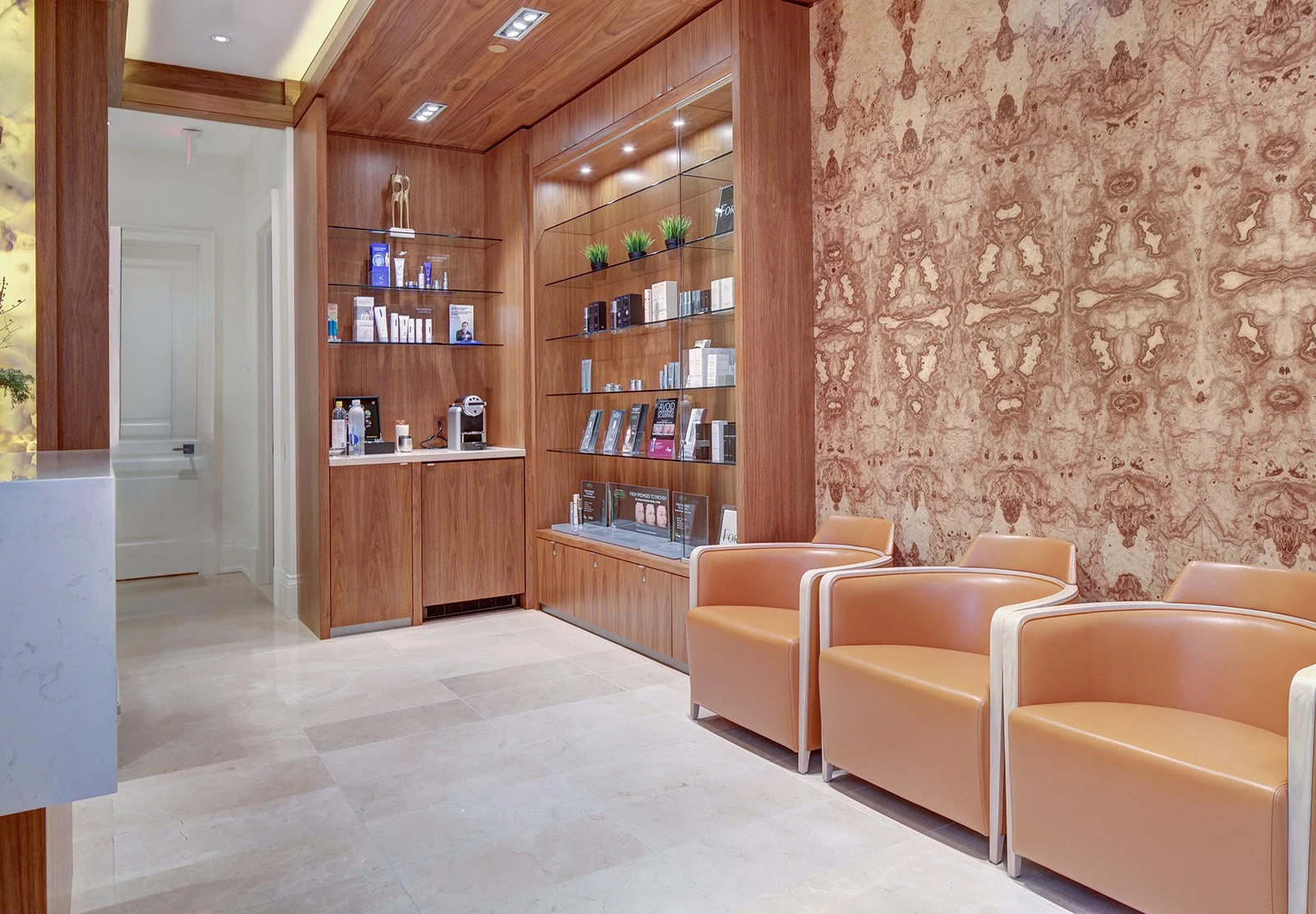 Waiting area with beige leather chairs, a wall with marble-like pattern, wooden shelves displaying skincare products, and a coffee station.