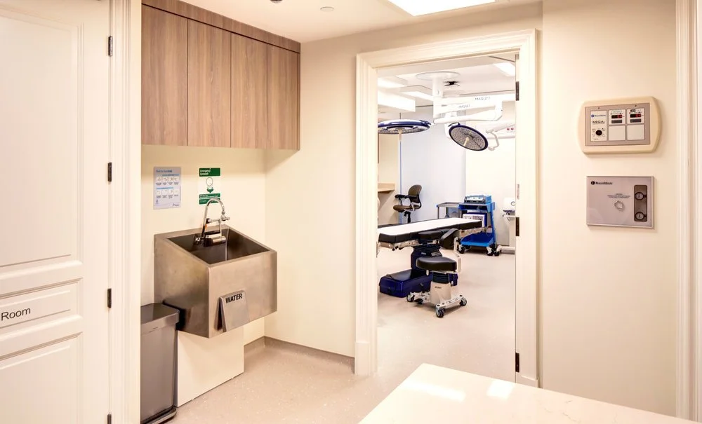 View of a hospital room with a medical examination table, surgical lights, and medical equipment, seen through an open doorway from a hallway with a water sink and control panel.