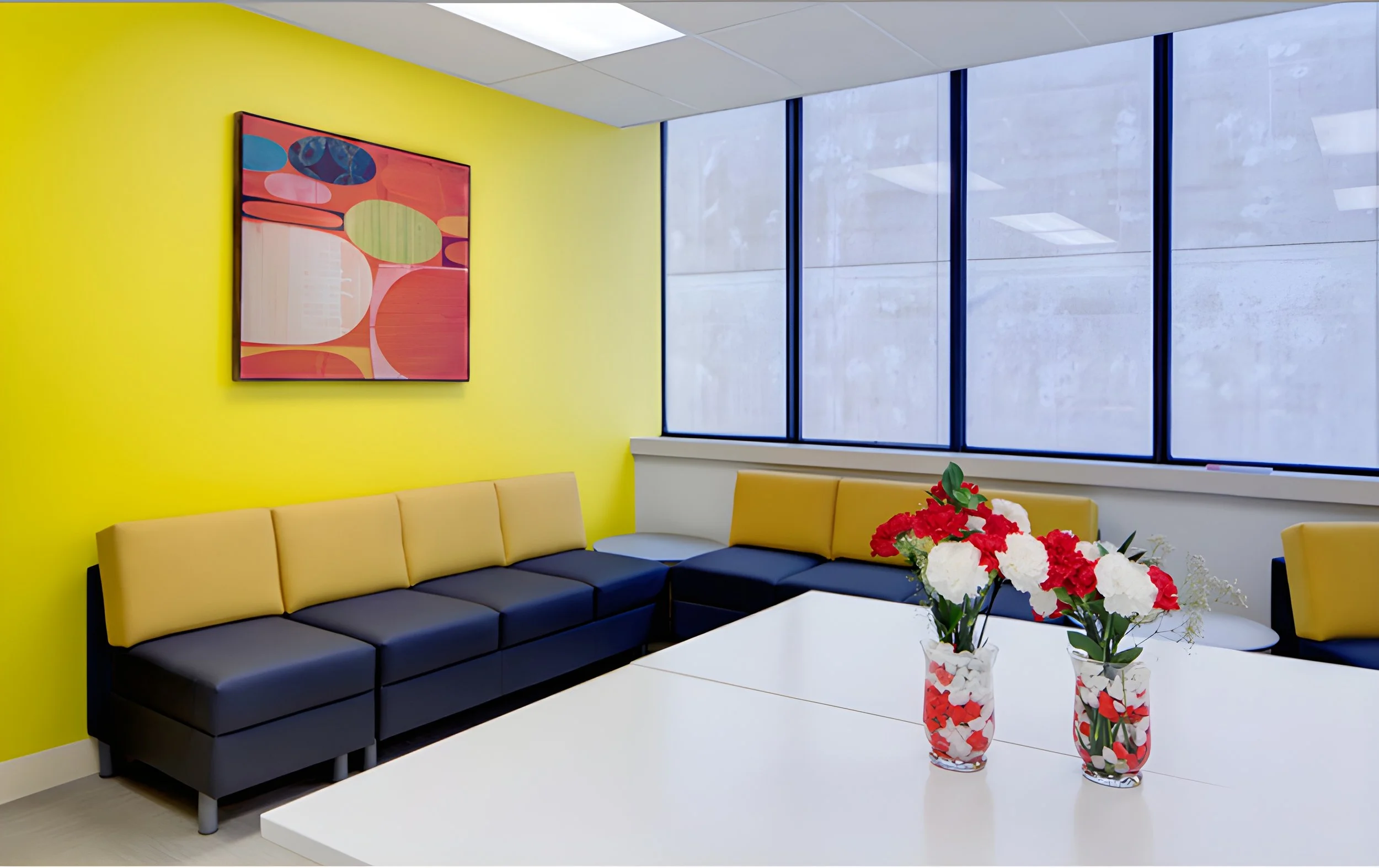Interior of a modern office lounge area with a yellow wall, a colorful abstract painting, a yellow sectional sofa, a white table with red, white, and pink flower vases, large windows with a concrete building exterior visible outside.