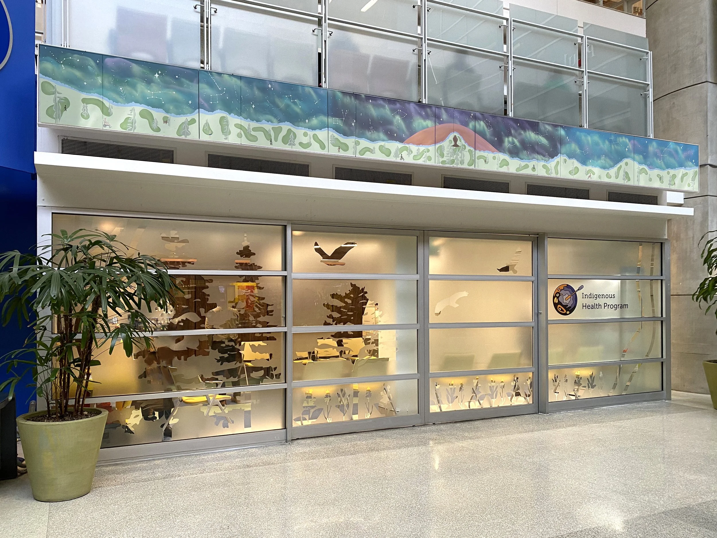 Indoor view of a building with large windows and a colorful mural of a mountain, trees, animals, and the night sky on the upper facade. There is a potted plant in the foreground and a sign reading 'Indigenous Health Program' on the glass below.