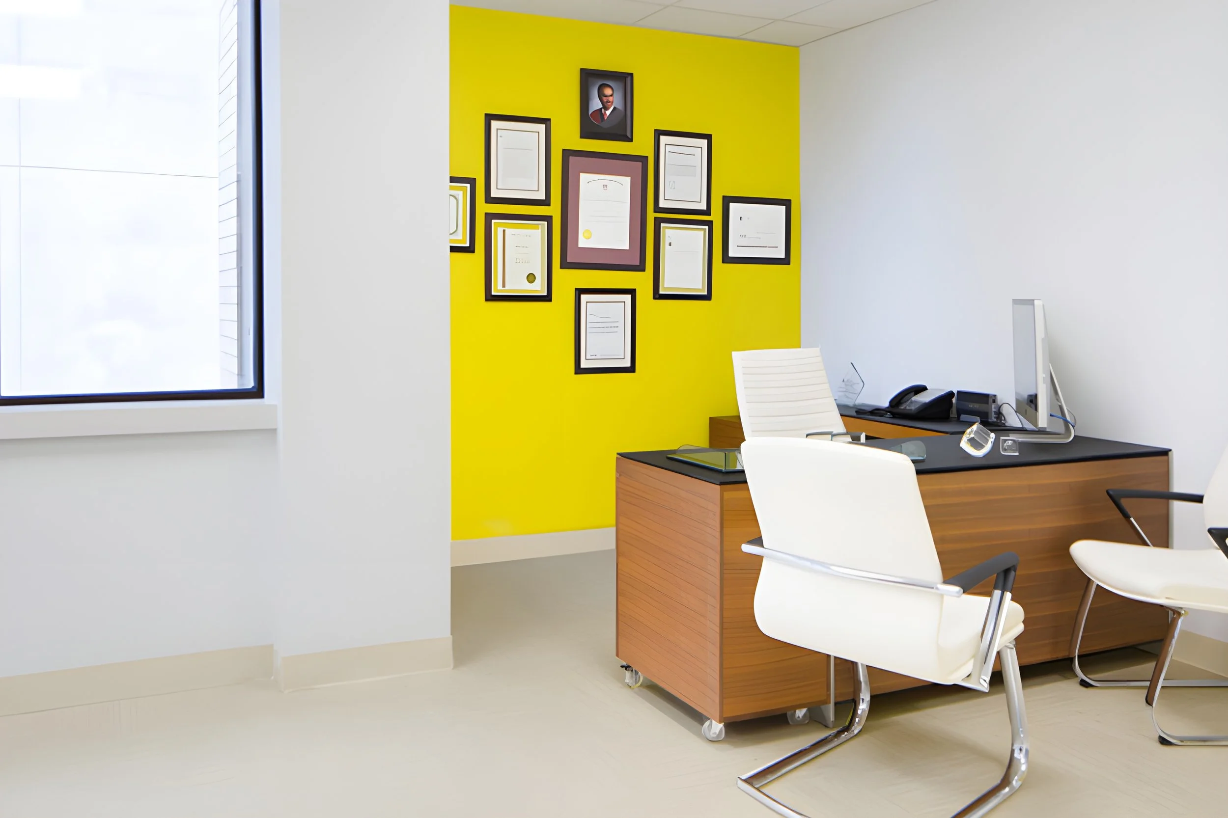 An office with a yellow accent wall decorated with framed documents and a portrait. The office has a large window on the left, a wooden desk with a white chair, a computer monitor, a telephone, and a second chair to the right.