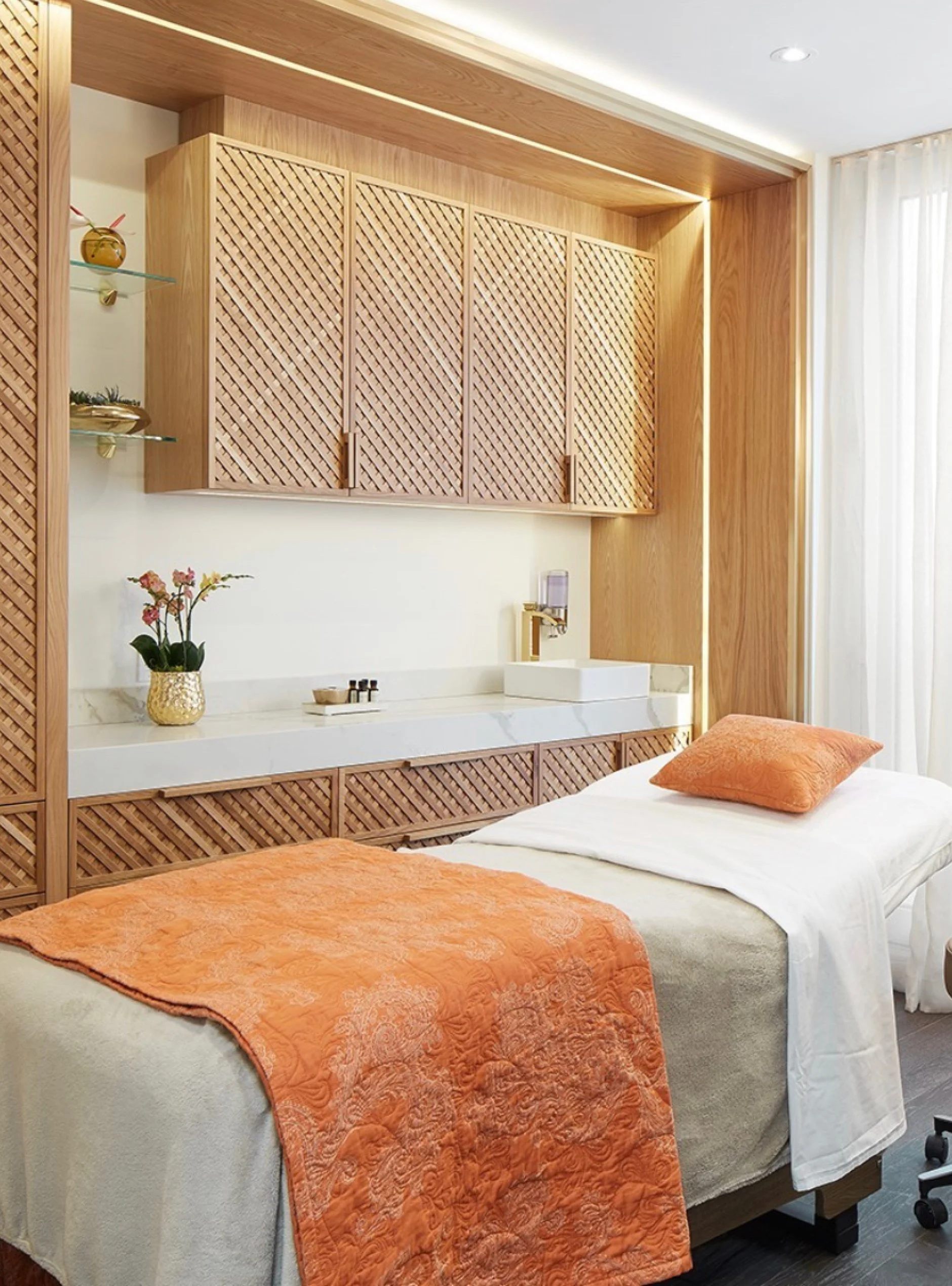 A bedroom featuring two beds with orange quilts and matching pillows, white sheets, a marble countertop with a plant and toiletries, wooden cabinets, glass shelves with decorative items, cream curtains, and modern lighting.