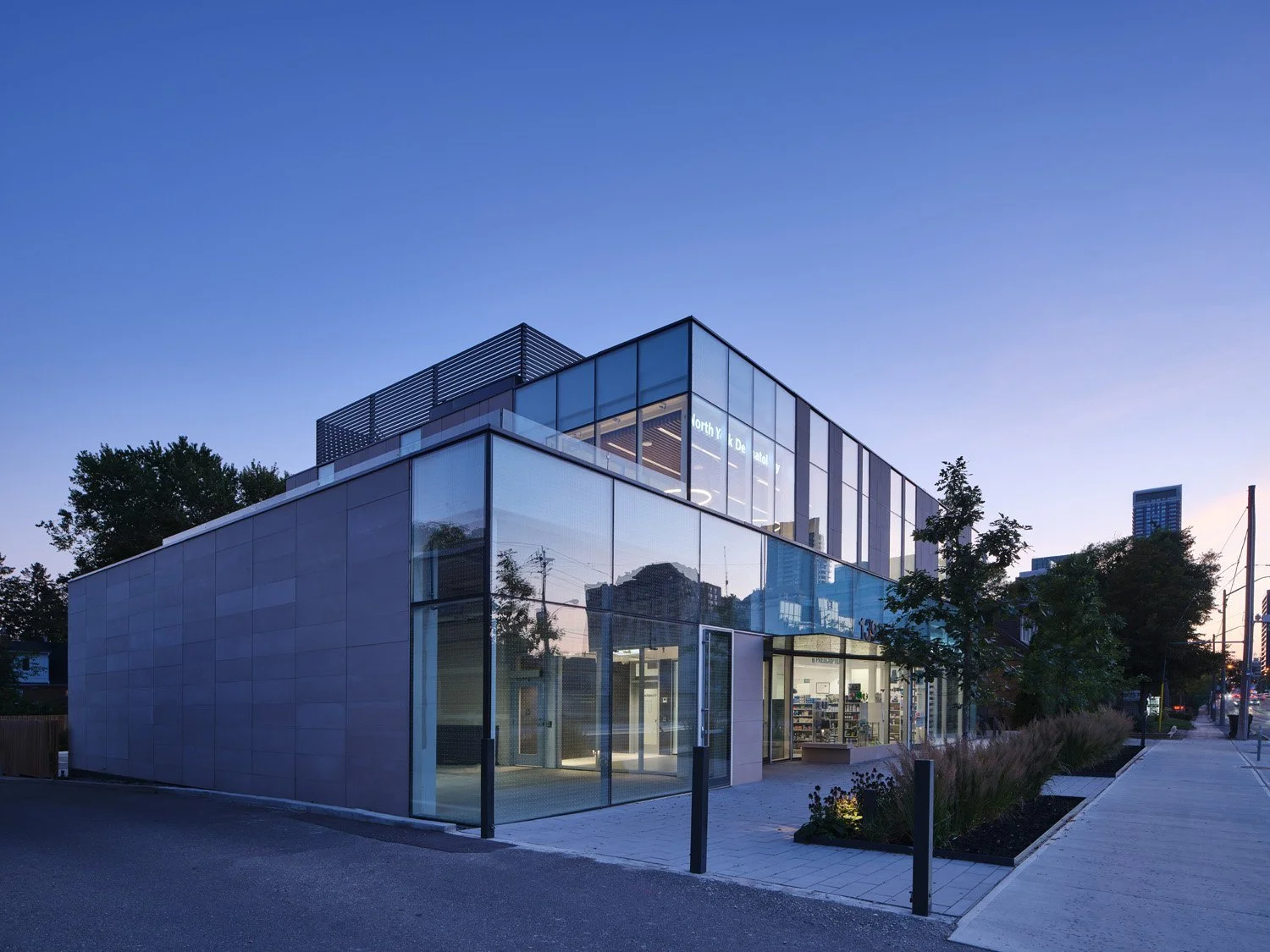 Modern two-story glass building during dusk with interior lights on, trees and cityscape in the background, and a sidewalk with landscaping in the foreground. Salutogenic design. Healthcare architecture. Inspiration for Toronto, Dallas and Park City.