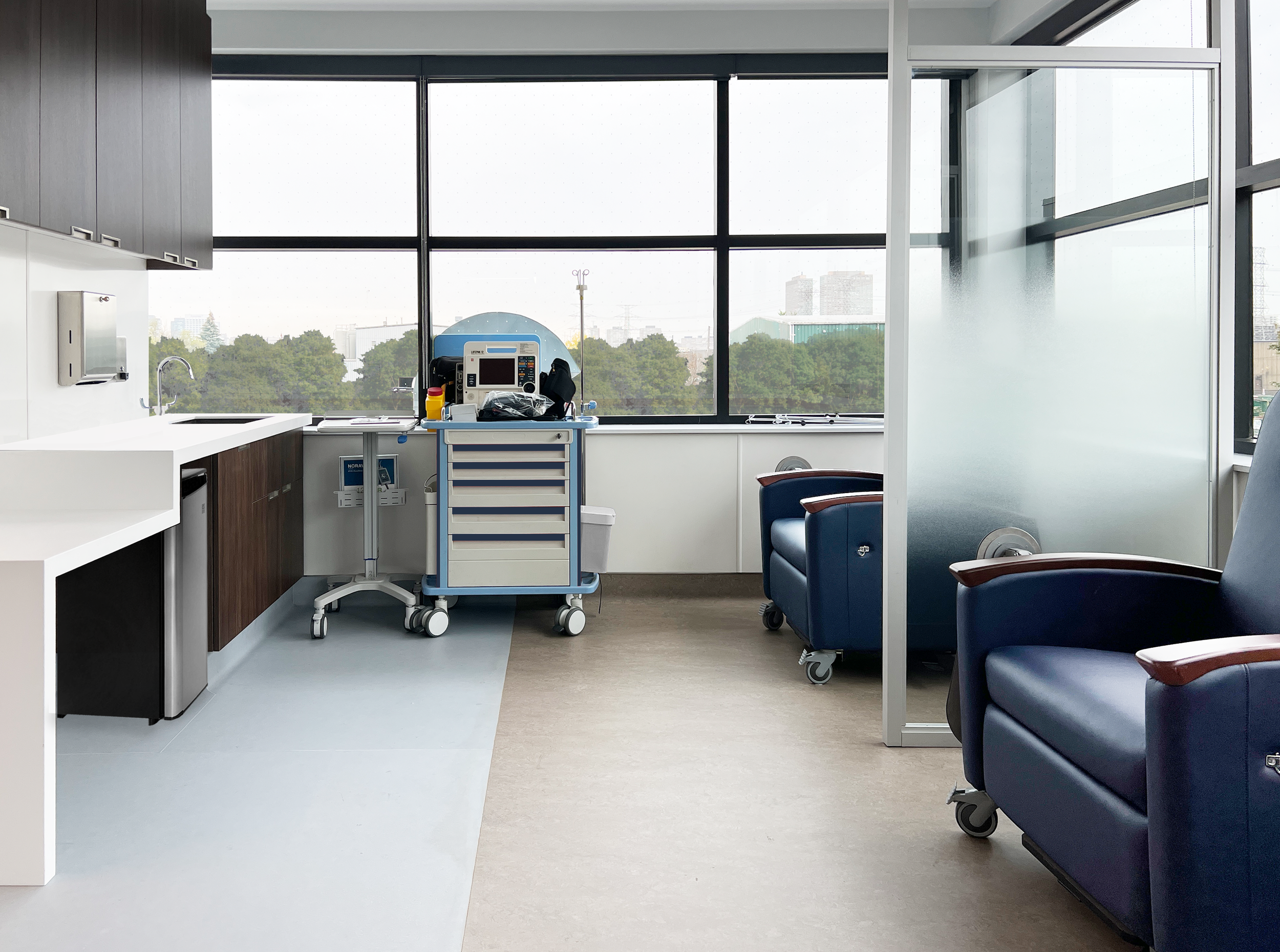 Medical examination room in a clinic in Toronto with chairs, medical equipment, and large window overlooking cityscape. Inspiration for Miami, Park City, Dallas and Texas. Salutogenic design, human flow, beautiful architecture, human design.