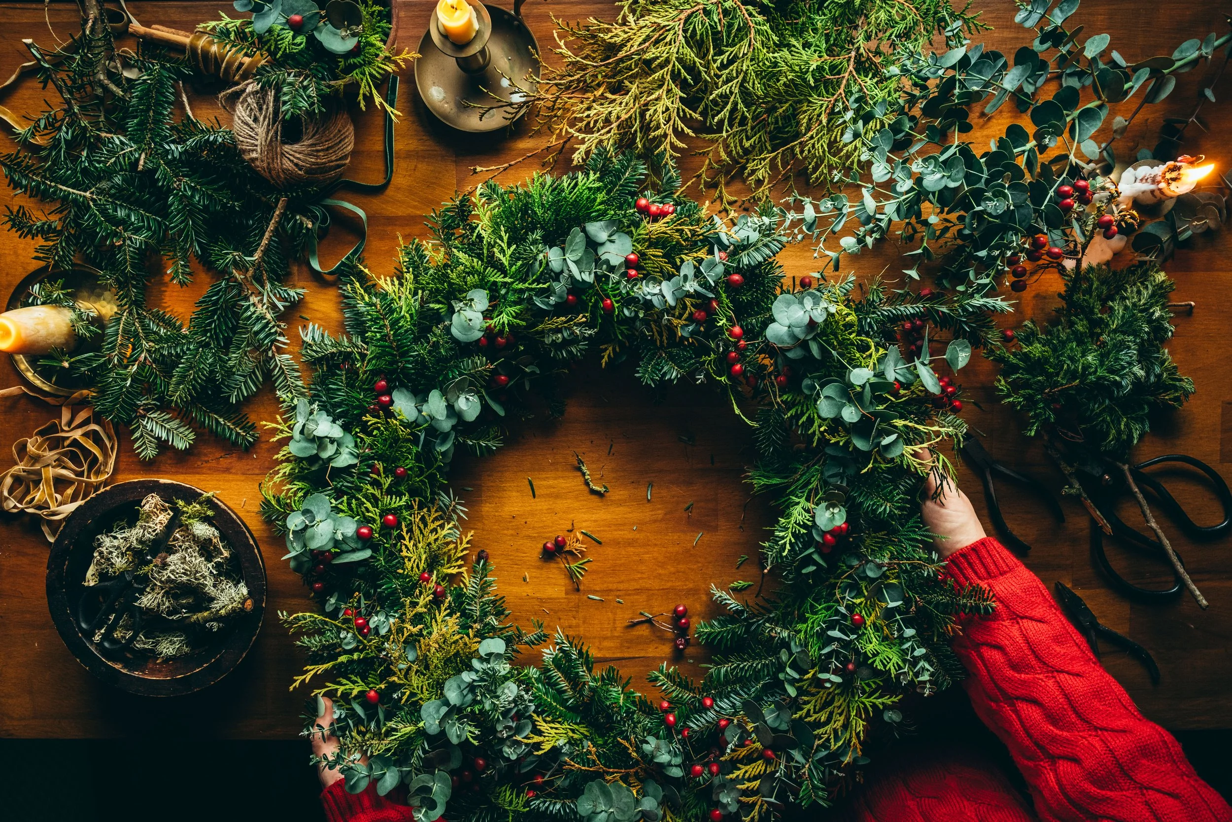A creative flatlay photograph of a hand made Christmas wreath made with fir tree branches, eucalyptus, foraged evergreens and hawthorn branches. The wreath is being held by a person wearing a knitted red woollen jumper which adds to the festive feel