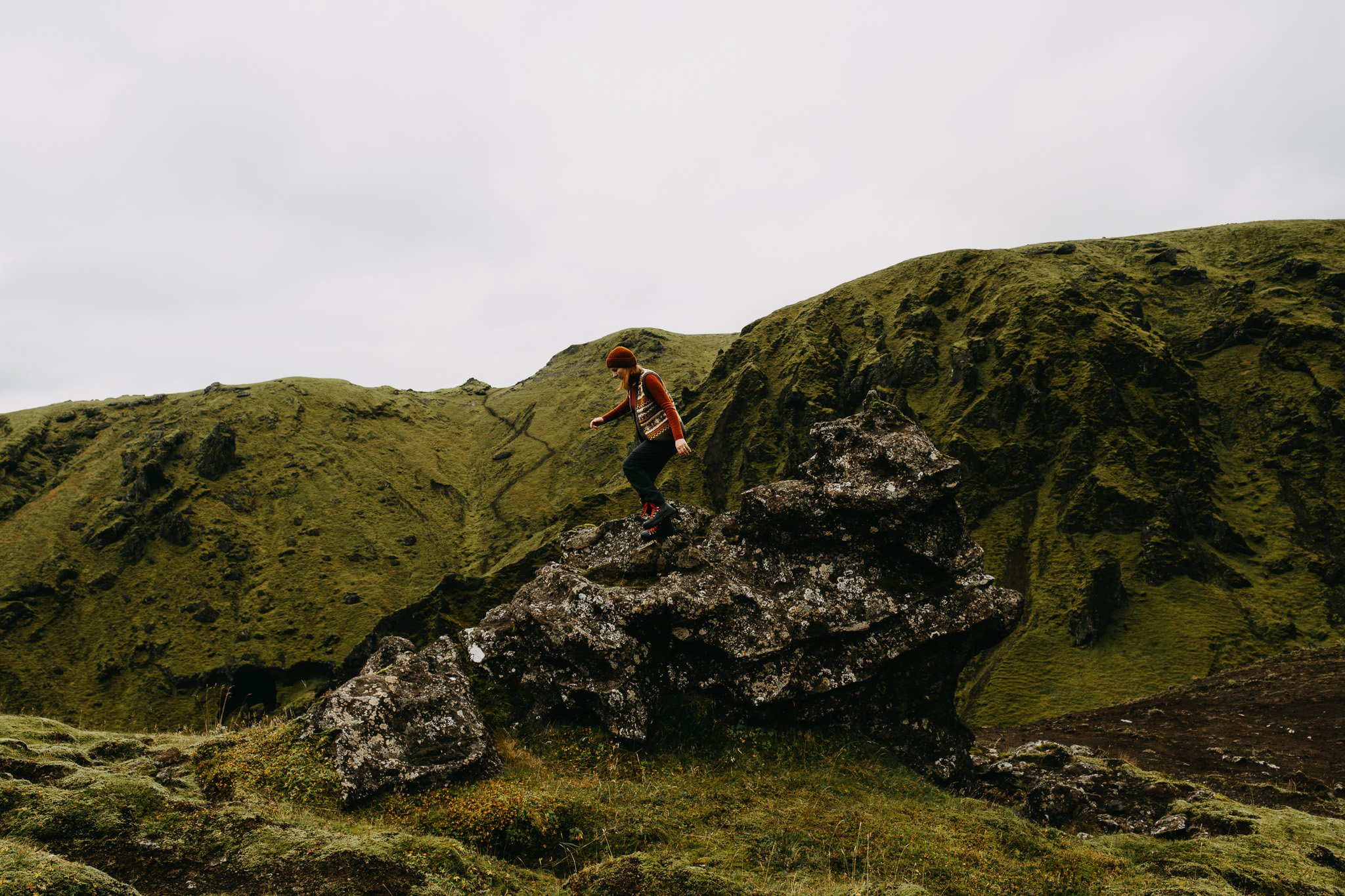 A woman wearing a sienna blouse and a fair isle vest climbs on a rock formation set in a green Icelandic mountain landscape