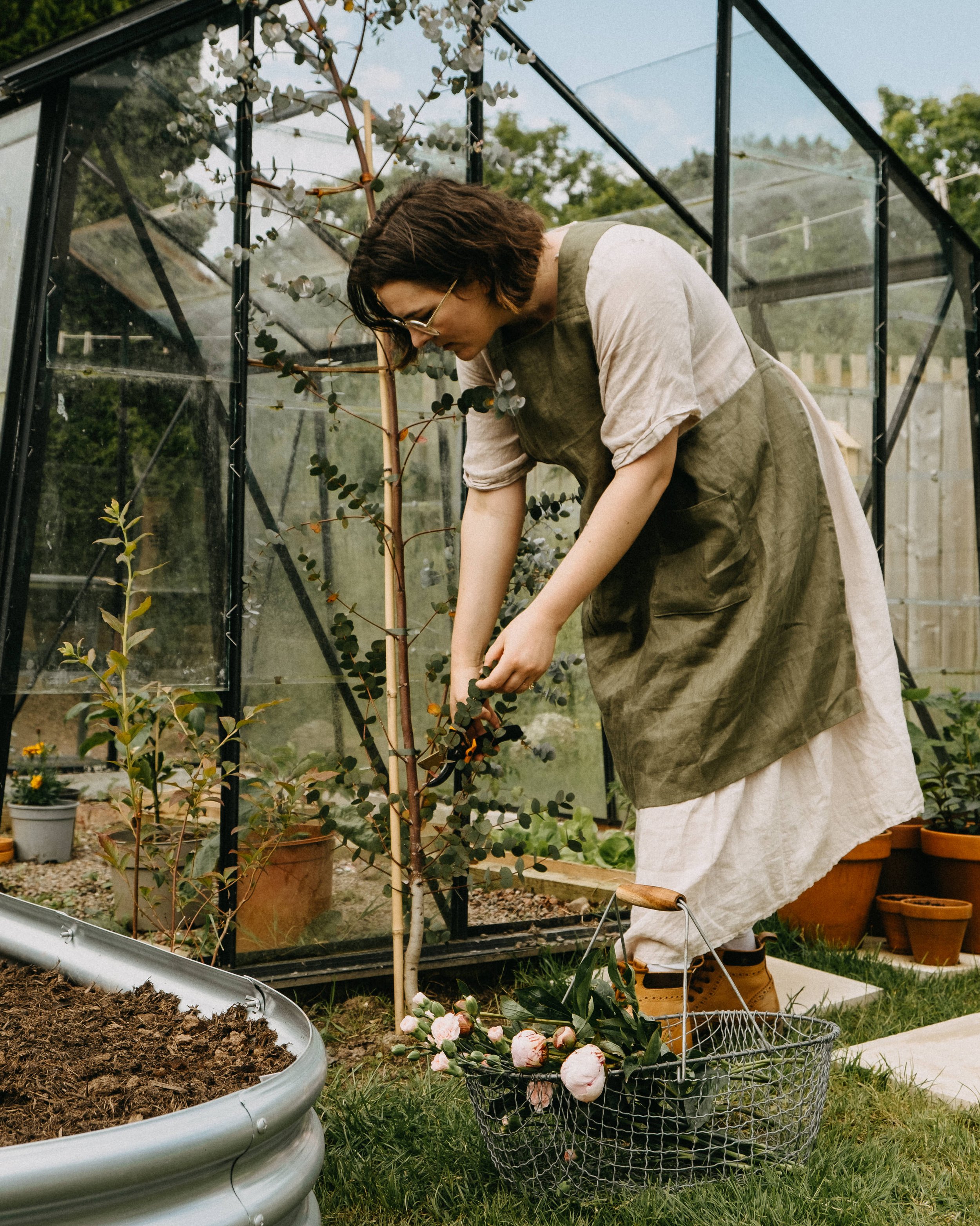 Self portrait photography, cottagecore outfit, wearing a linen dress with pinafore apron in front of the greenhouse.