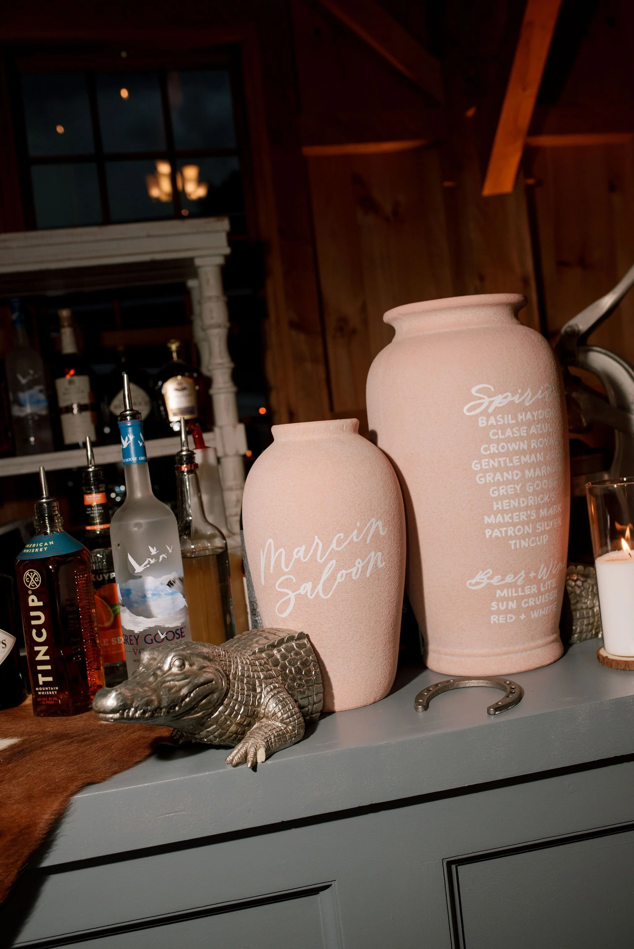 Decorative bar with two pink vases, a small crocodile figurine, bottles of alcohol, and bar tools on a counter, at an Orange County wedding.