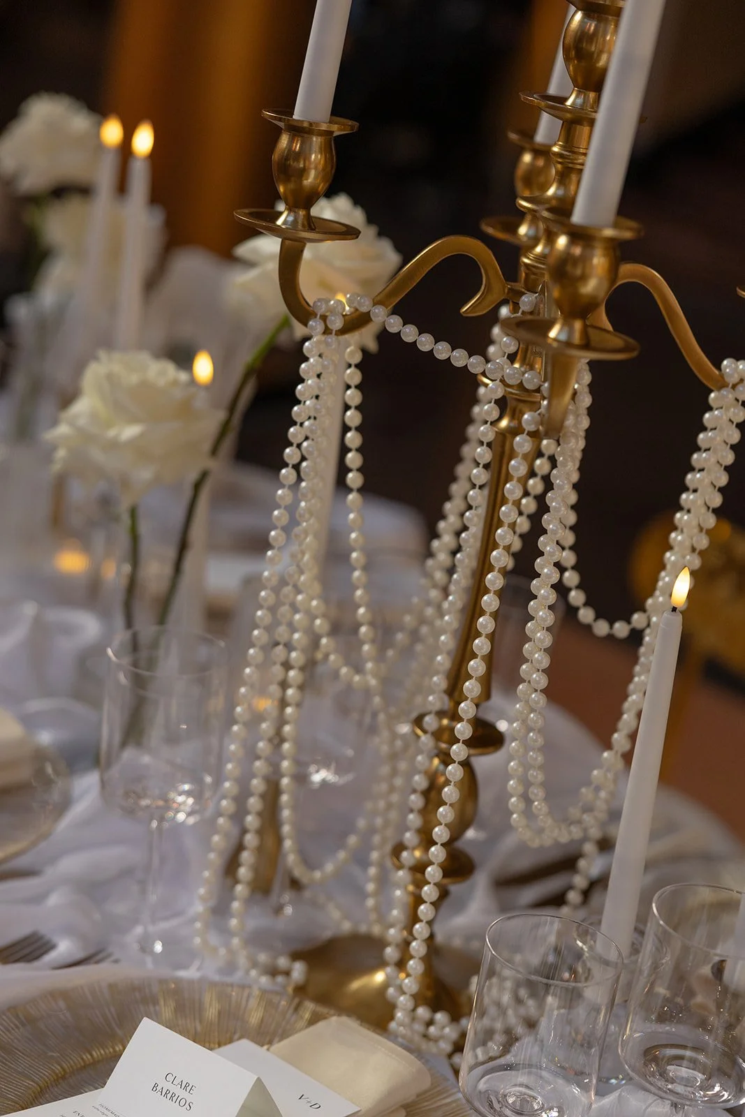 Close-up of a gold candelabrum adorned with pearl beads, with white candles and white roses in a luxurious wedding table setting.
