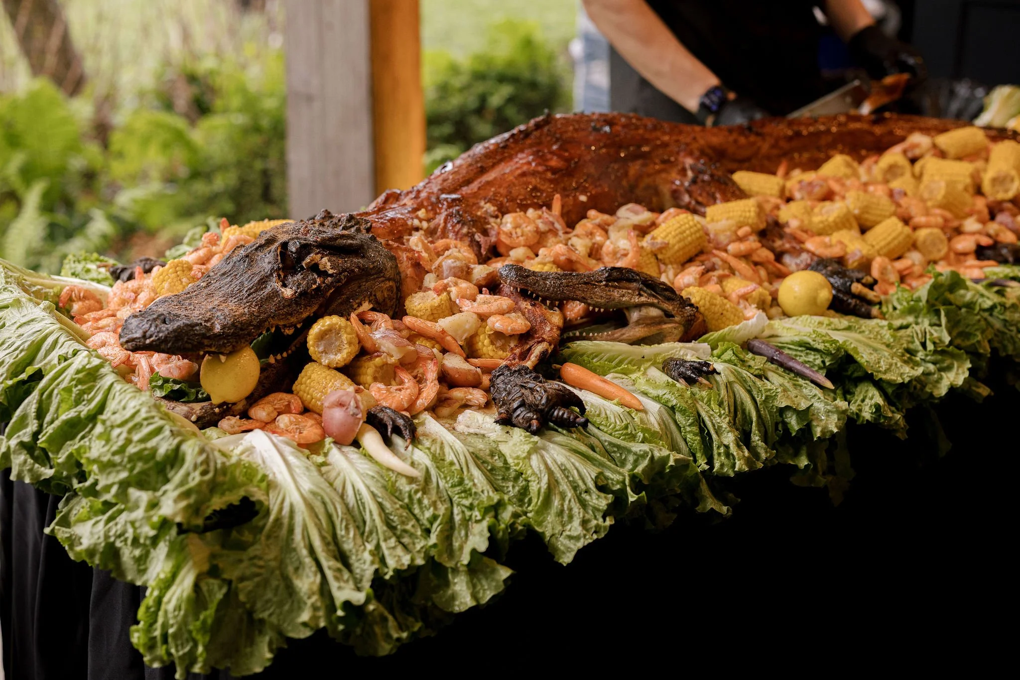 A cooked aligator laying on a bed of lettuce, garnished with vegetables and fruits, outdoors on a table.