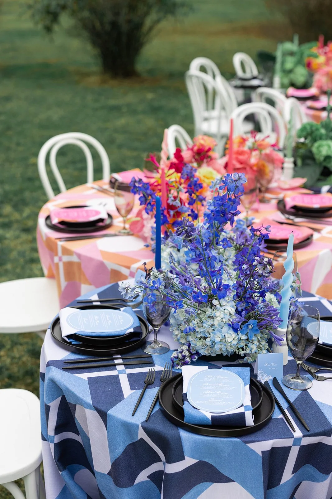 A colorful outdoor wedding or event table setup with a floral centerpiece, candles, plates, glasses, and cutlery, arranged on a patterned tablecloth surrounded by white chairs.