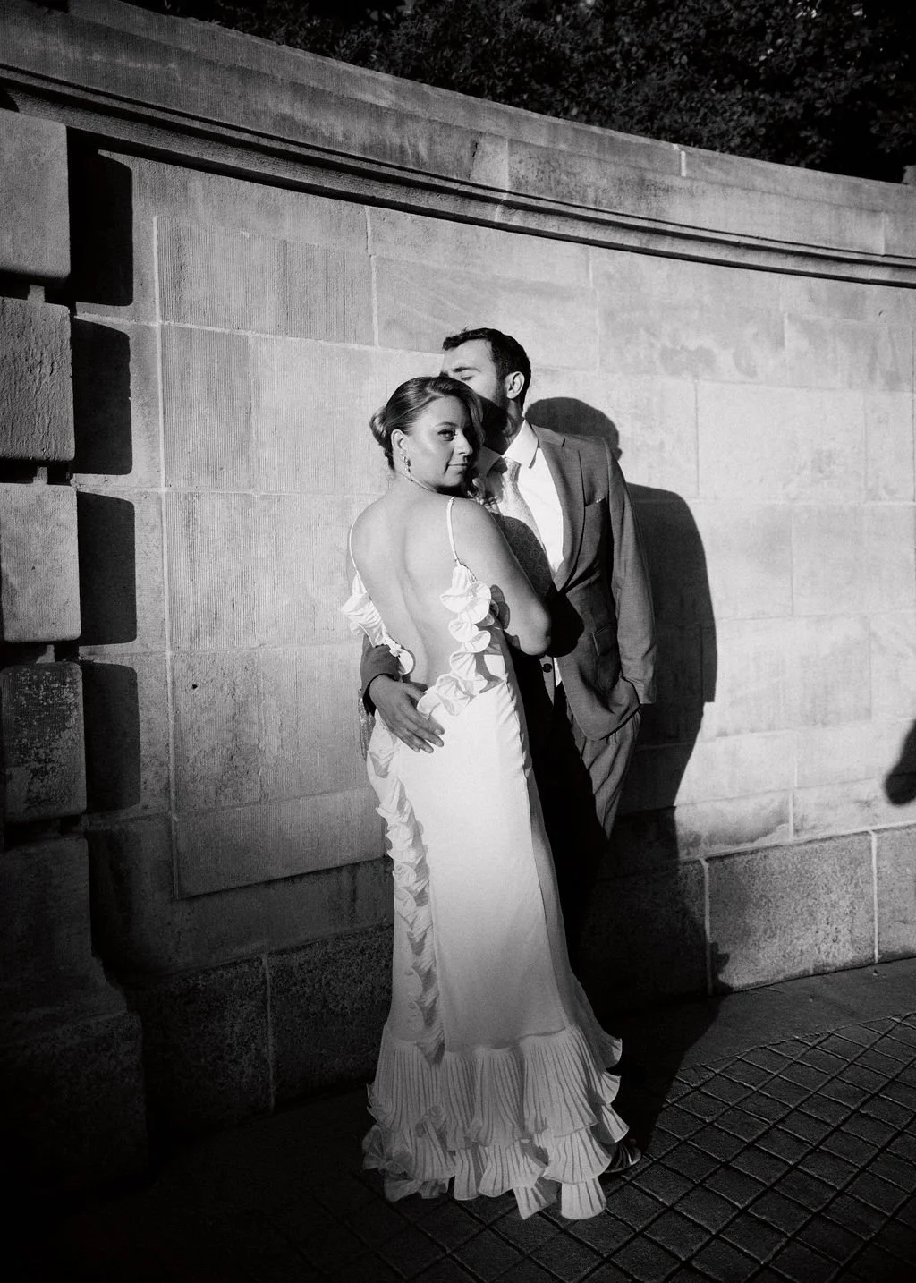 A black and white photo of a couple at night, with the woman wearing a fancy dress and the man in a suit, standing close against a stone wall.