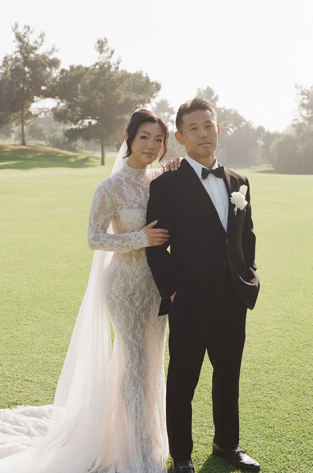 A bride and groom standing on a green lawn during daytime, with trees in the background.