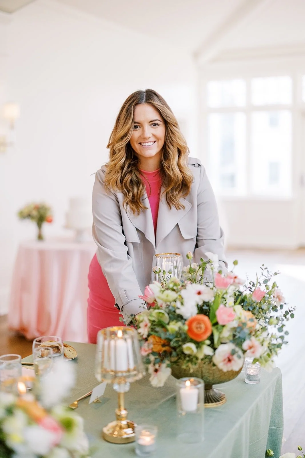 Lizzie Noble, creative director of Lizzie Noble Events, smiling at a wedding reception table with a floral centerpiece, candles, and glasses, in a bright room.