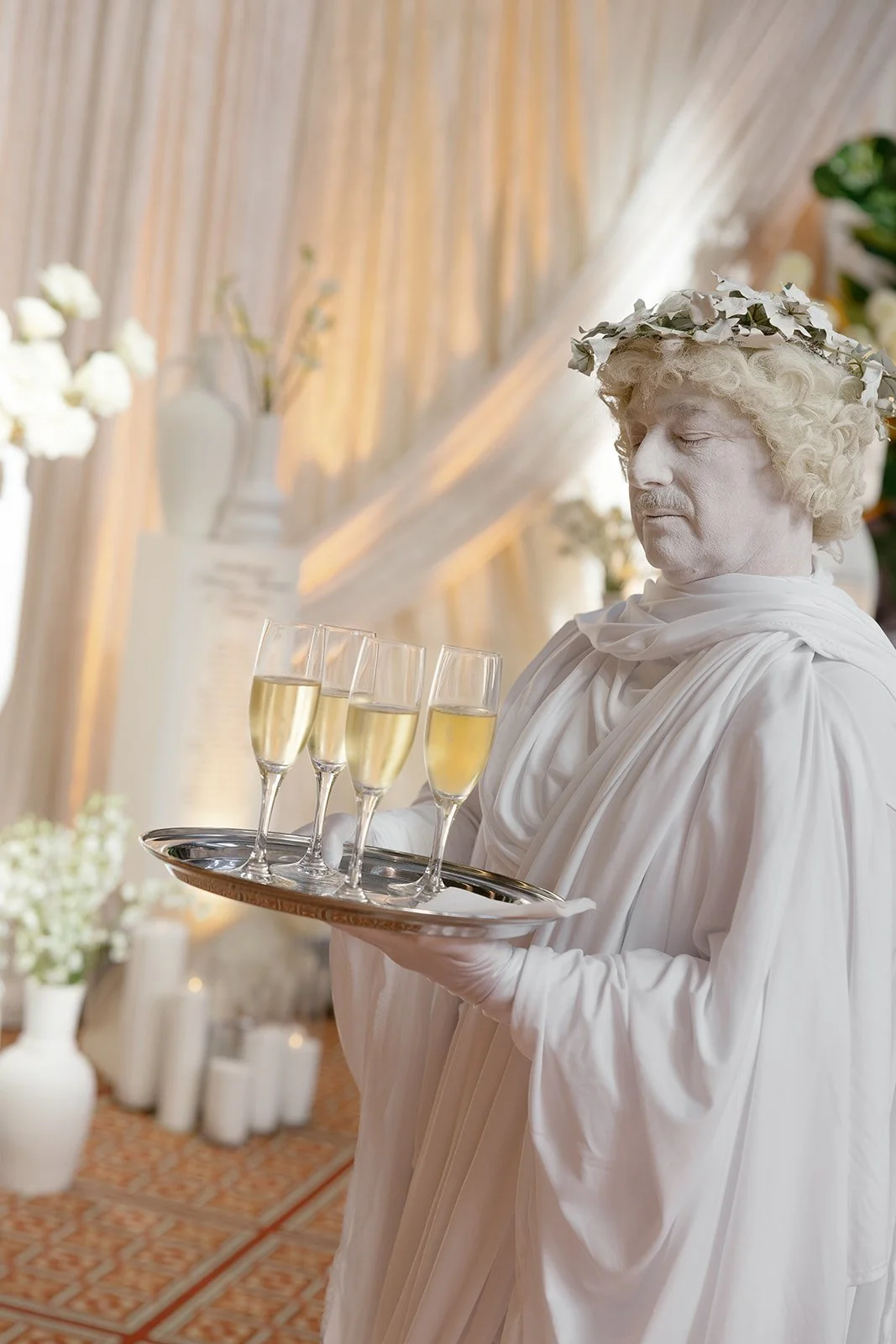 A person dressed as a Greek god, with a white toga, curly blonde wig, and a leafy crown, holding a tray of four glasses of champagne at a luxury Orange County wedding.
