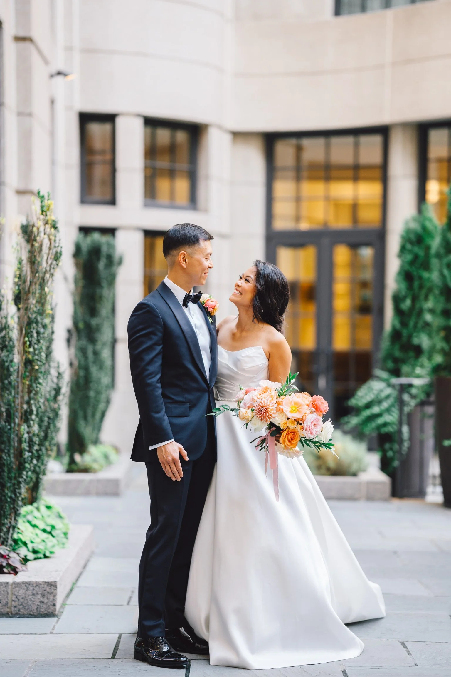 A newlywed couple smiling at each other during their wedding in a California outdoor setting with modern building architecture, greenery, and large windows.