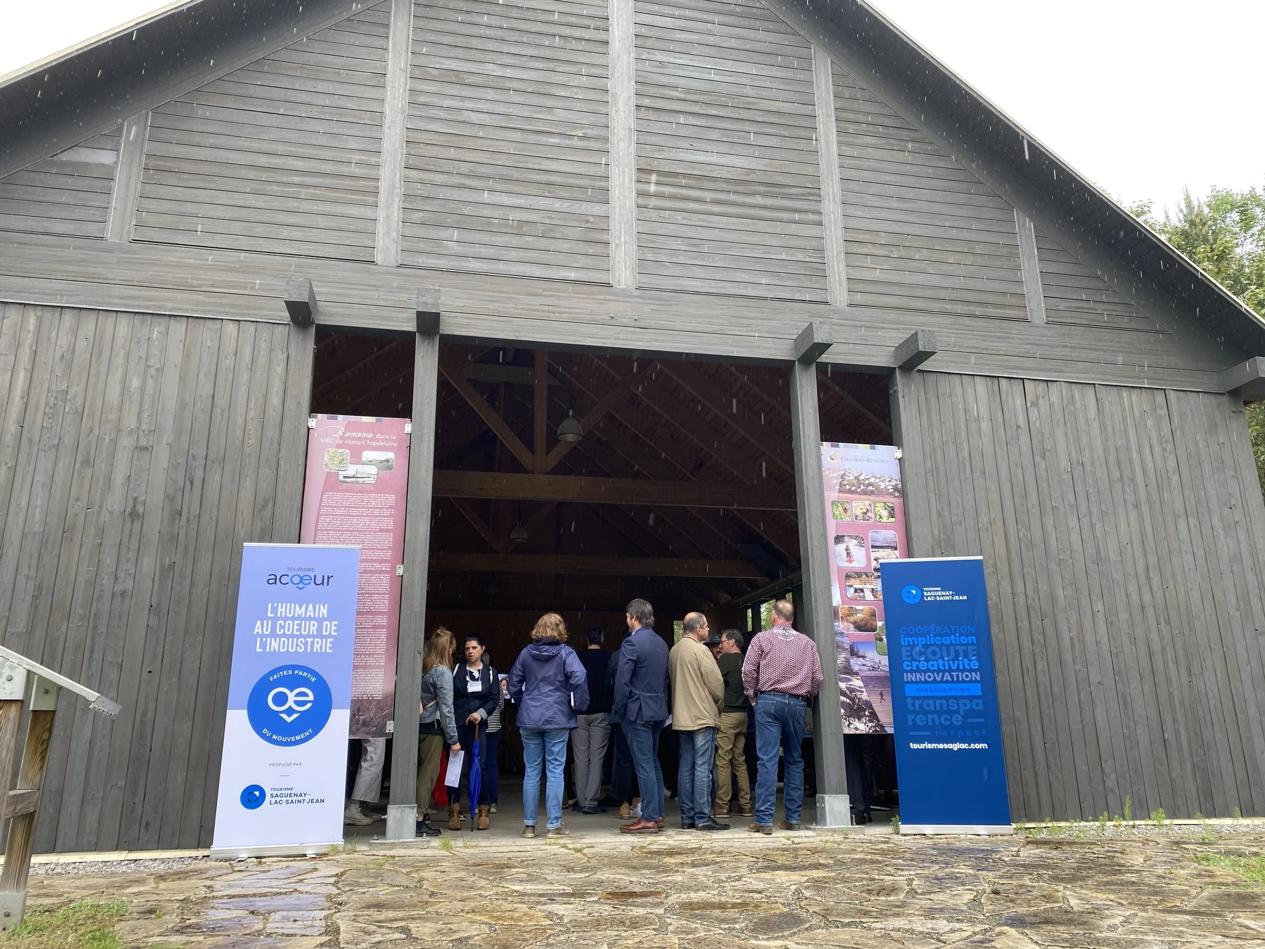 Groupe de personnes devant un bâtiment en bois lors d'une réunion ou d'un événement, avec des affiches promotionnelles à l'entrée.