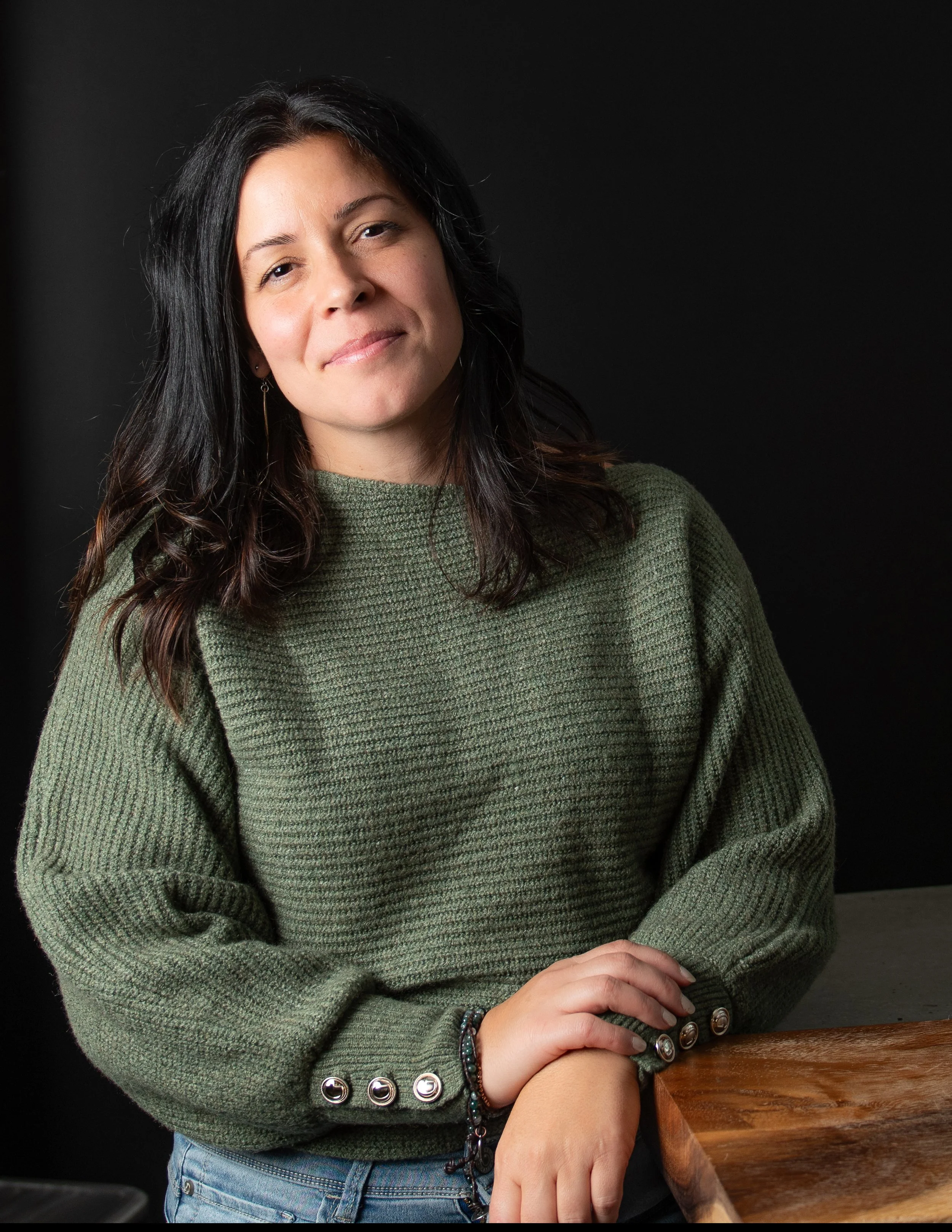 Jeune femme aux cheveux longs noirs portant un pull vert en gros plan sur fond noir.