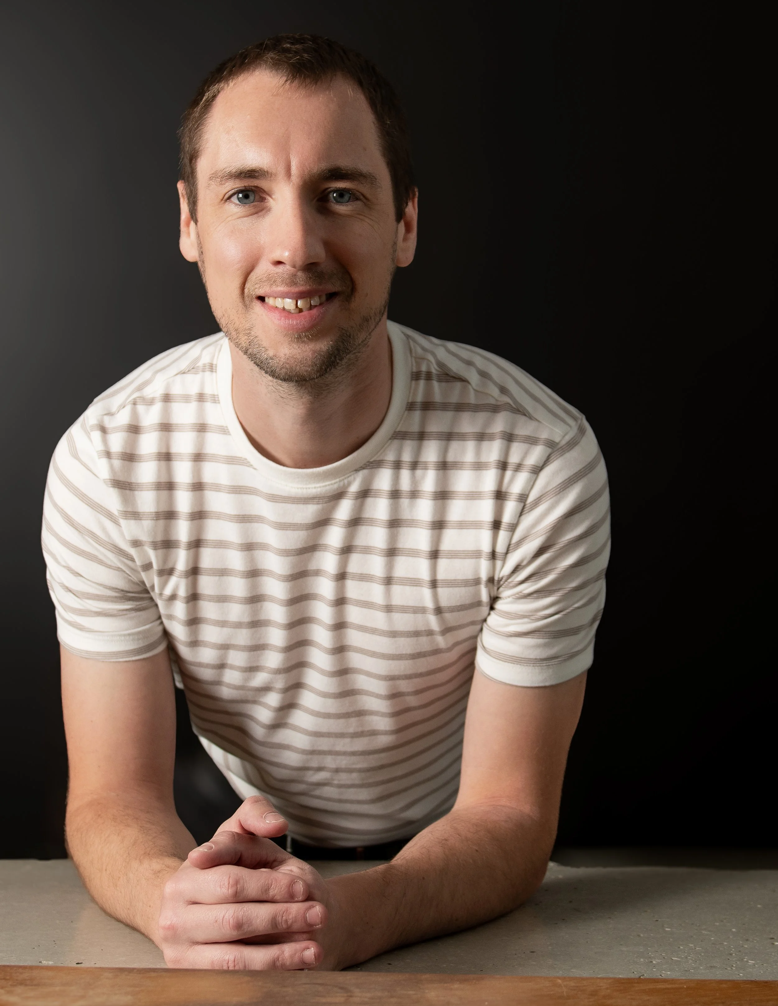 Un jeune homme souriant devant un fond noir, portant un t-shirt blanc à rayures
