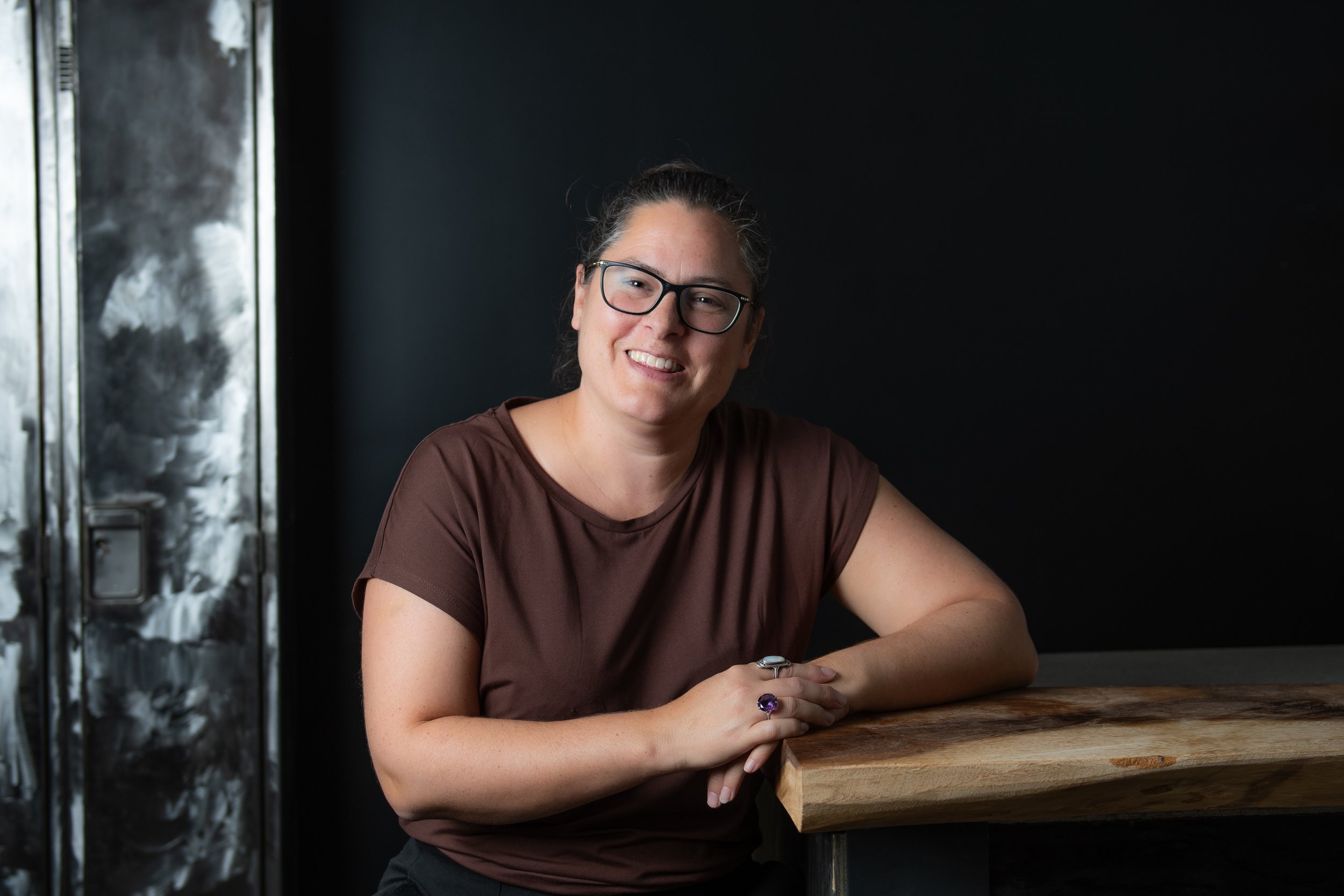 Femme souriante avec lunettes, portant un t-shirt marron, assise à une table en bois, dans un environnement sombre.