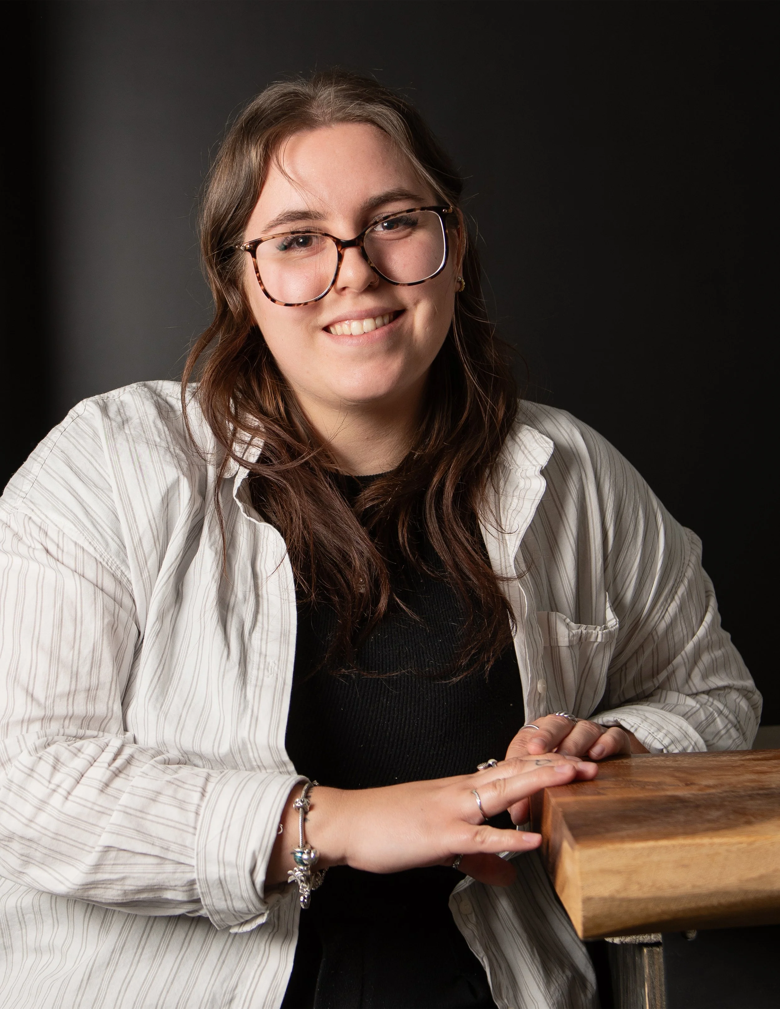 Jeune femme souriante portant des lunettes, tenant une planche en bois, contre un fond sombre.