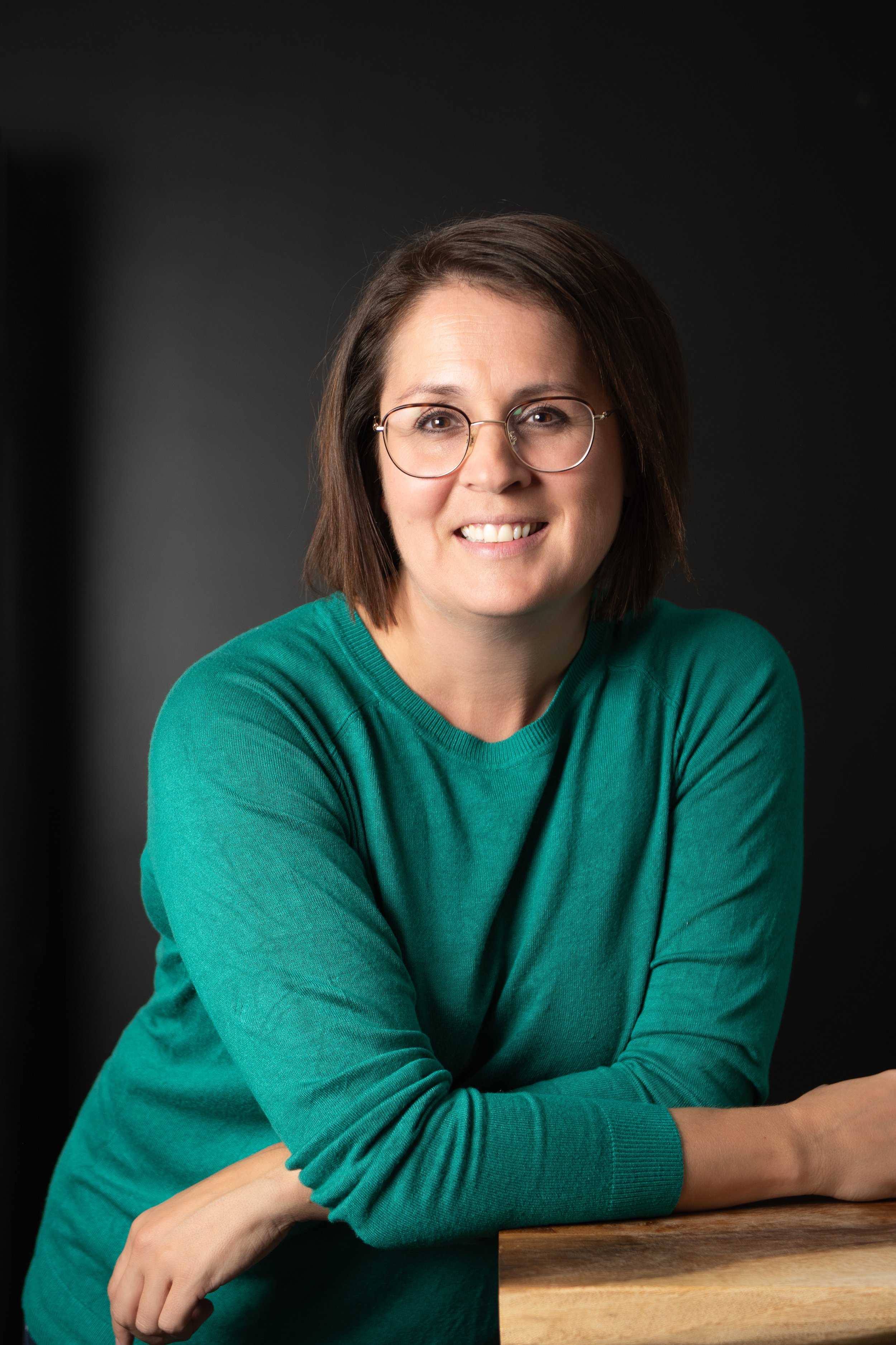 Une femme portant des lunettes, souriante, avec une chemise verte, posant devant un fond noir