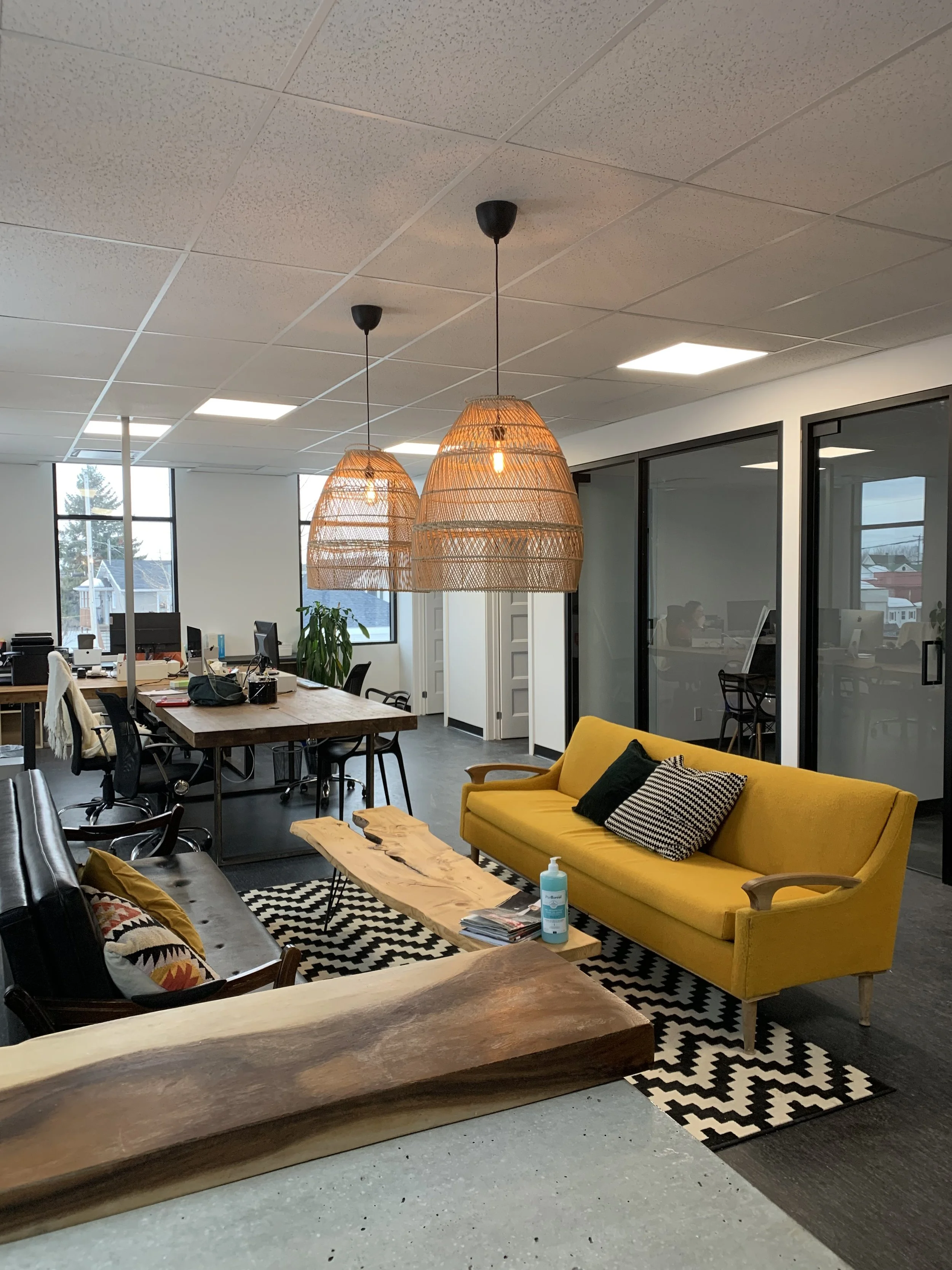 Un espace de bureau moderne avec un canapé jaune, un tapis noir et blanc, une table en bois, des lampes en rotin suspendues, des fenêtres donnant sur des bâtiments et des employés travaillant à des bureaux séparés par des murs en verre.