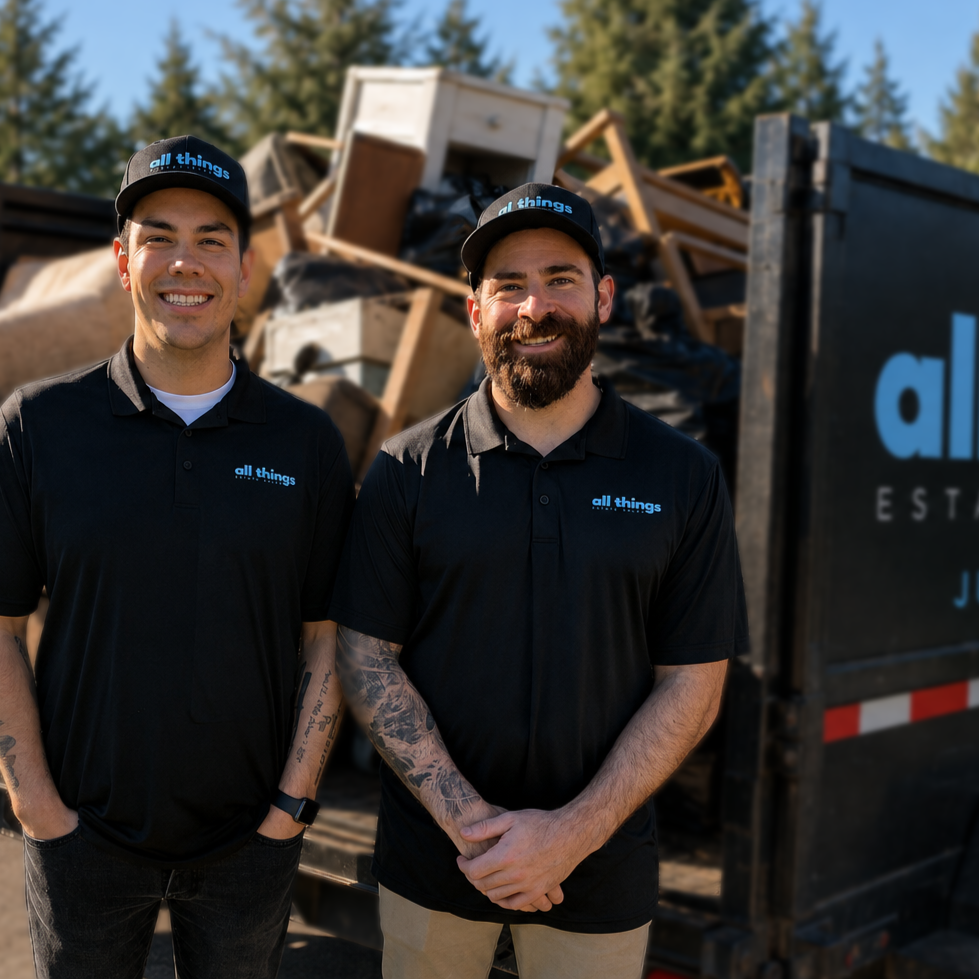 Two smiling men in black uniforms with "all things" logos on shirts and hats, standing in front of a truck filled with debris and wooden furniture outdoors on a sunny day.