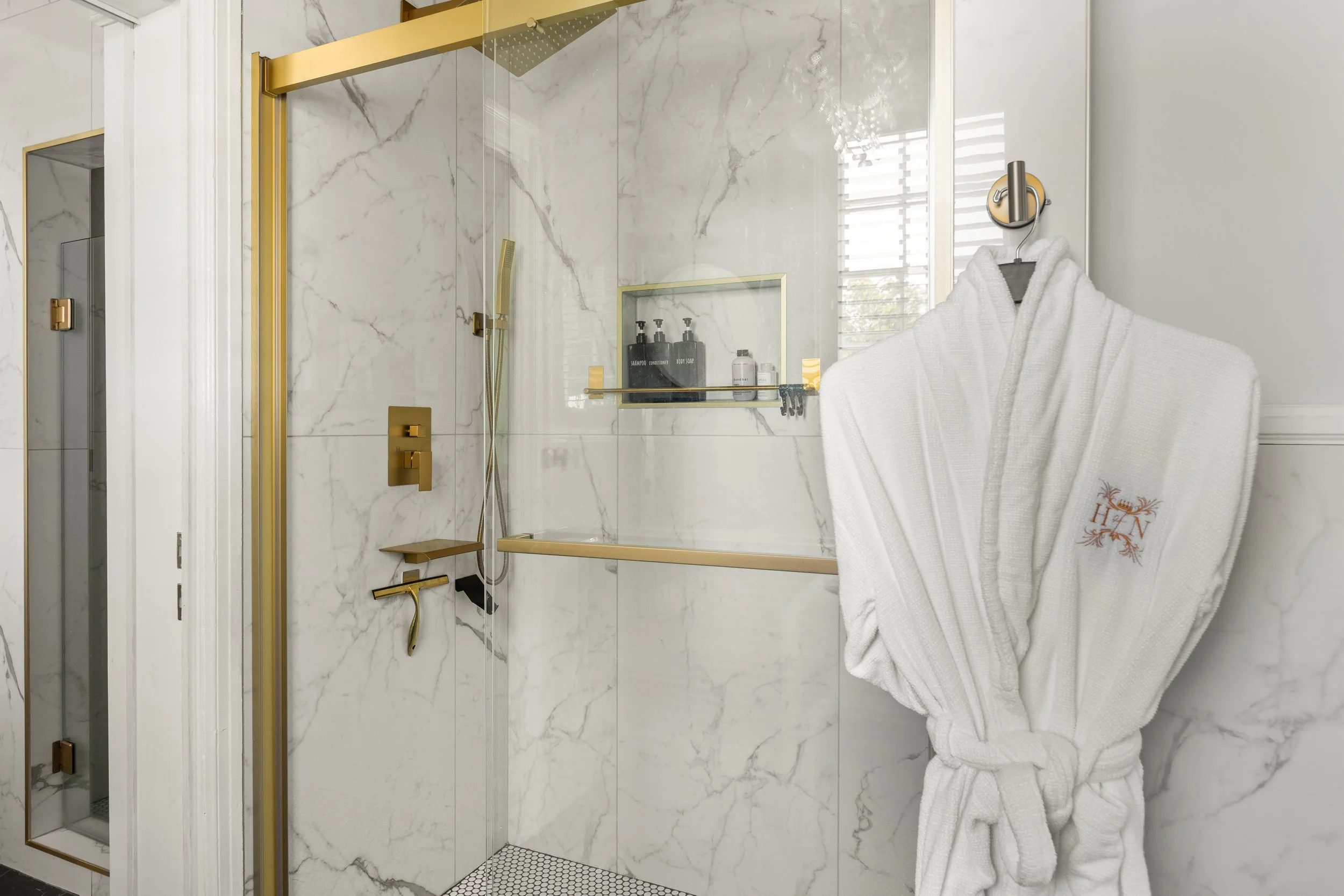 A luxurious bathroom shower area with white marble walls accented by gold fixtures, a glass door, and a white robe hanging on the wall with embroidered initials.