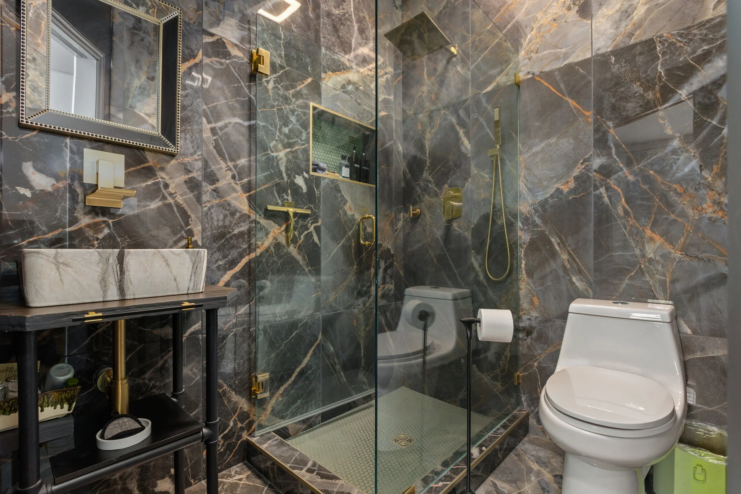 A luxurious bathroom with marble walls and floor, a glass shower enclosure with gold fixtures, a white toilet, a black vanity table with a marble basin sink, and a mirror with a decorative gold frame.