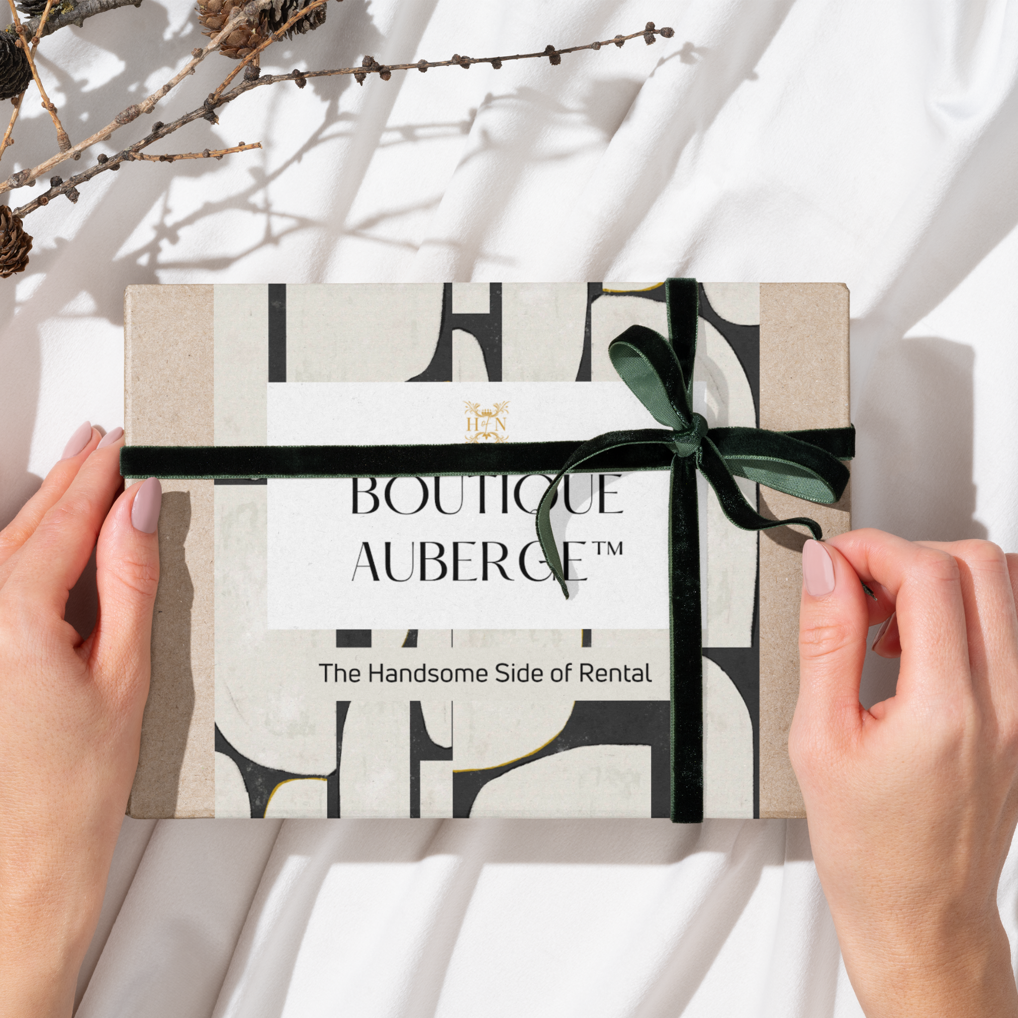 A person holding a rectangular gift box with a black ribbon tied in a bow on top, resting on a white cloth surface with dry twigs nearby.