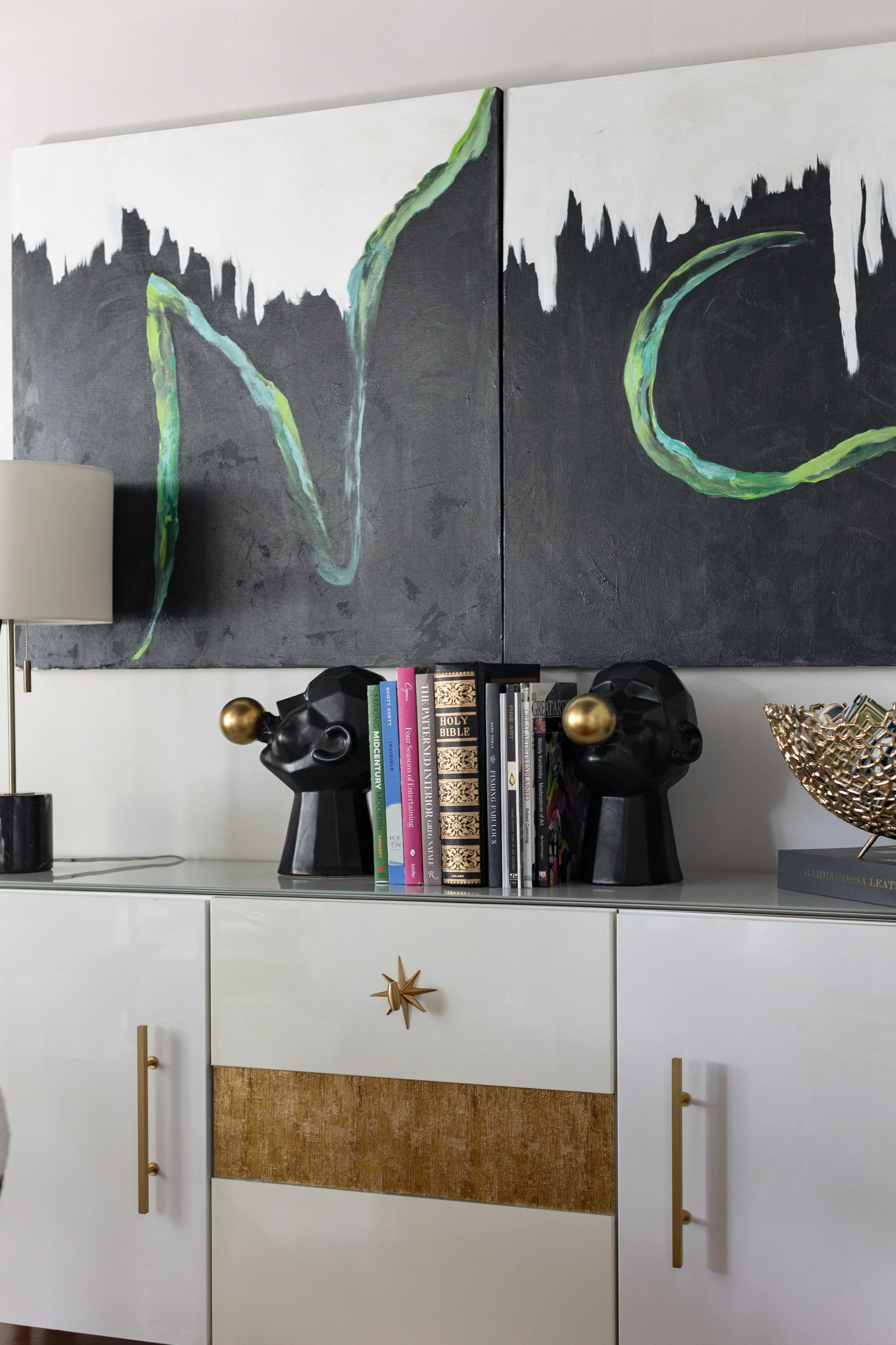Close-up of a modern white console table decorated with two black sculptural head vases with gold spheres, a row of colorful books, a gold decorative bowl, and a large abstract black and green painting hanging above.