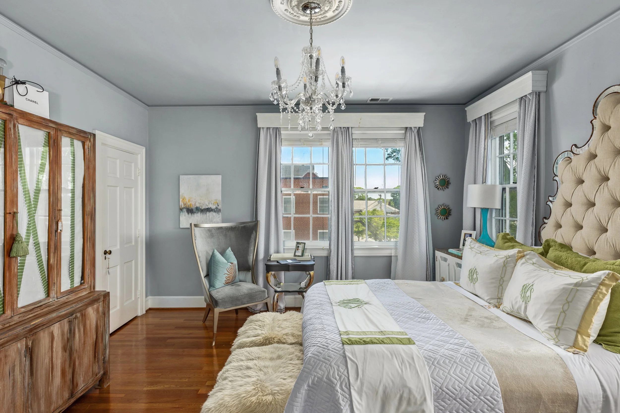 A bedroom with light gray walls, a large beige tufted headboard, and white bedding with green accents. There are three windows with white curtains, a chandelier hanging from the ceiling, and a wooden wardrobe on the left. A small framed art piece hangs on the wall, and there's a gray armchair near the window with pillows. A side table with a blue lamp and framed photo is next to the bed.