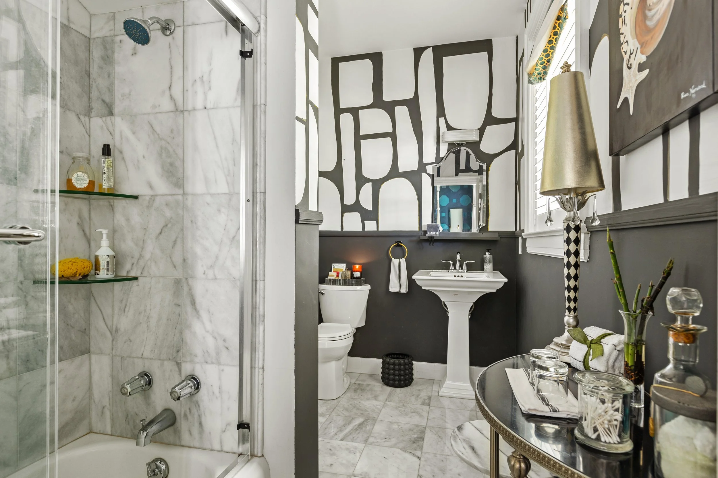 Bathroom with a glass-enclosed shower on the left, a toilet, a pedestal sink, and a wall with black and white abstract patterned design. Decor includes a lamp, plants, and various toiletries on a countertop and table.