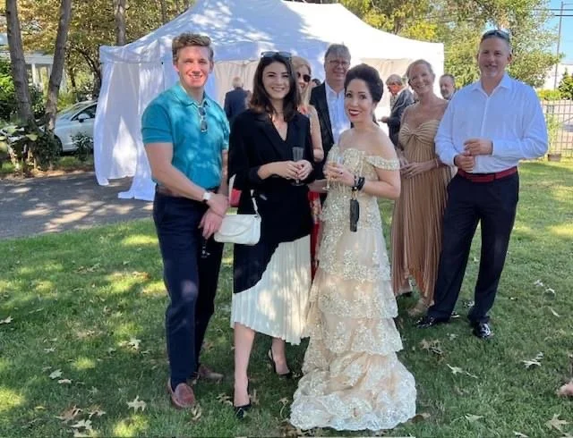 A group of seven people smiling outdoors at a celebration under a white tent, including a woman in a wedding dress and others in semi-formal attire.