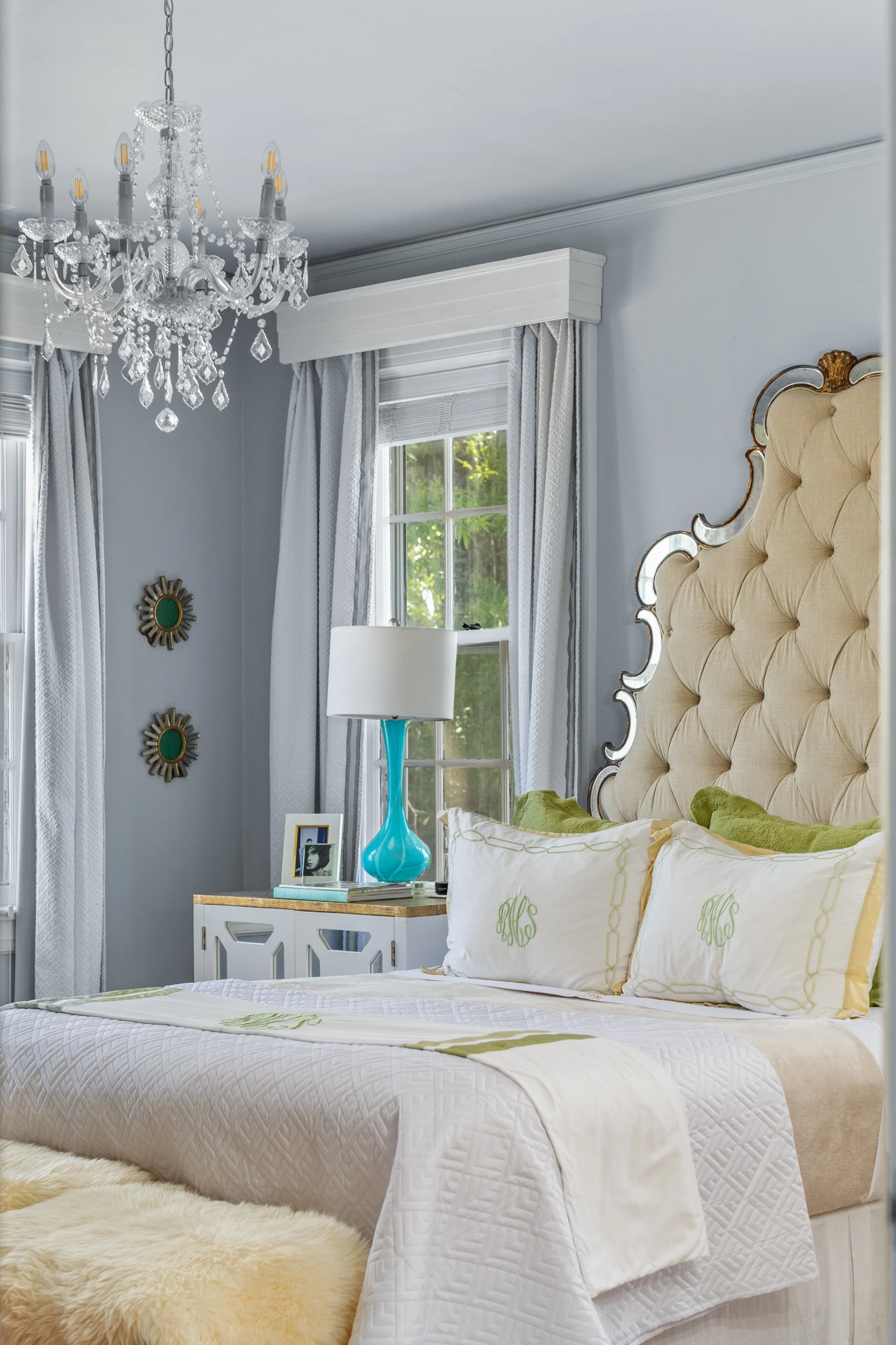 A bedroom with a large, tufted beige headboard, white bedding with monogrammed pillows, a white nightstand with a turquoise lamp, and a chandelier hanging from the ceiling. There are windows with white curtains and natural light.