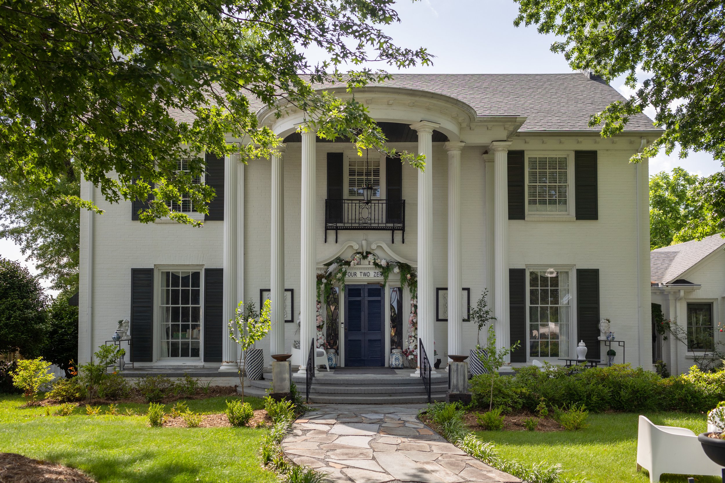 A large white house with black shutters and tall columns, decorated with floral arrangements and a sign that reads 'Four Two Zero,' surrounded by trees and a landscaped yard.