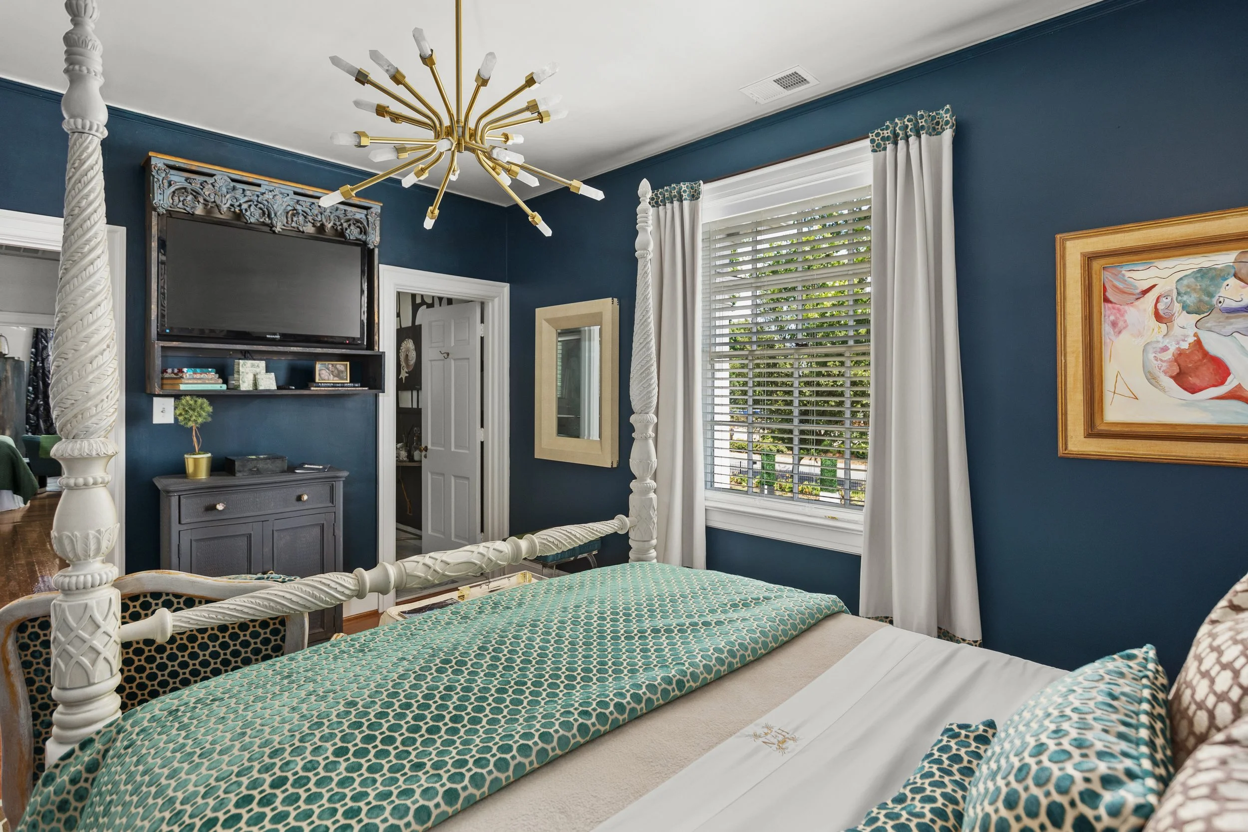 A bedroom with blue walls, a white four-poster bed with patterned bedding, a window with blinds and white curtains, a gold chandelier, a wall-mounted TV, and a framed abstract painting.