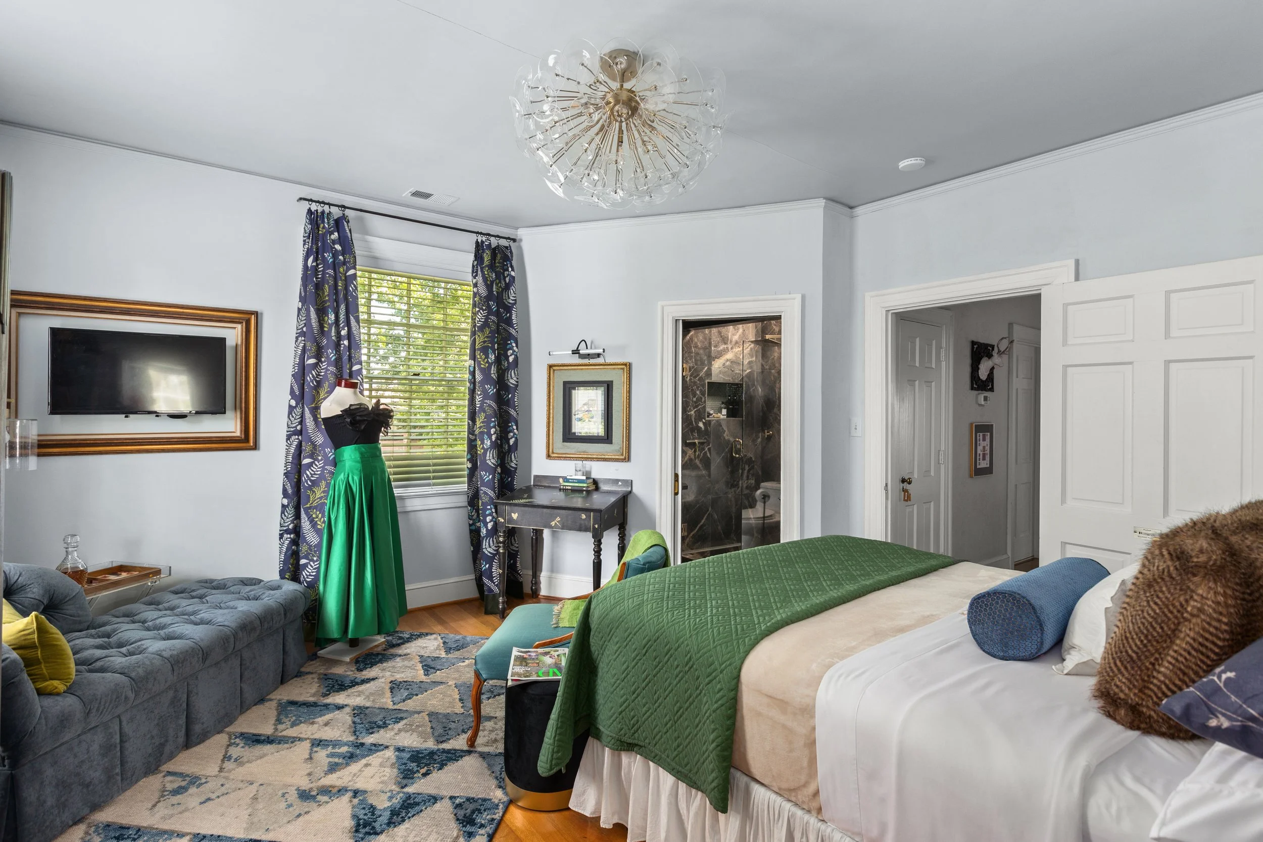 A bedroom with a bed, a velvet chaise lounge, a mannequin wearing a dress, a window with blue patterned curtains, a small desk, and a doorway leading to a bathroom.