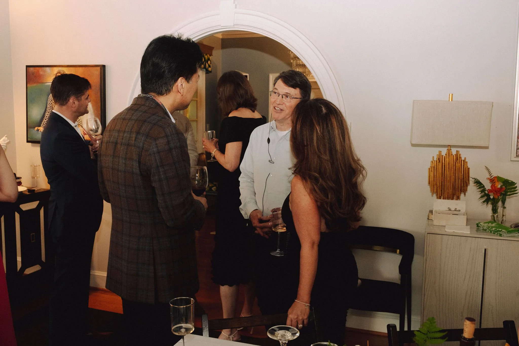 People socializing at an indoor party, holding wine glasses, in a well-decorated living room with artwork and a table with drinks.